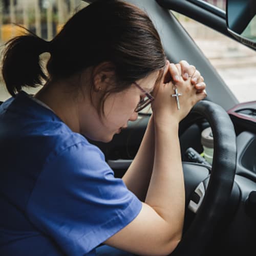 Female brunette nurse in the car praying with mask hanging from rearview mirror
