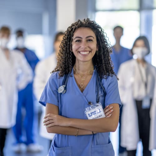 A large group of medical professionals stand in the hallway of a Hospital as they pose together for a portrait. They are each dressed professionally in scrubs and lab coats and some are wearing medical masks to protect them from airborne viruses.