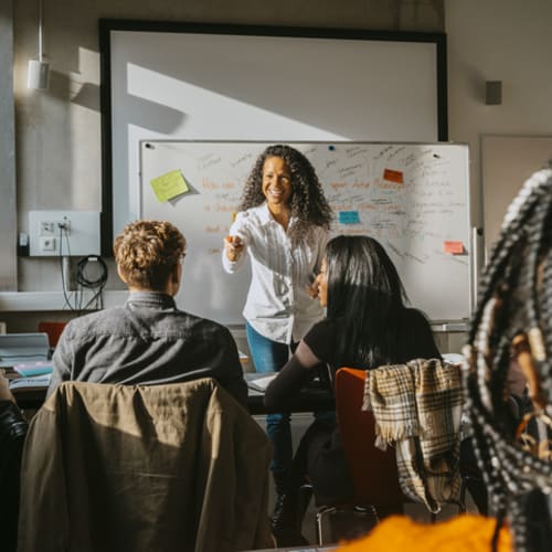 Brunette curly haired teacher stands in front of a white board engaging her students in breaking down a sentence's syntax