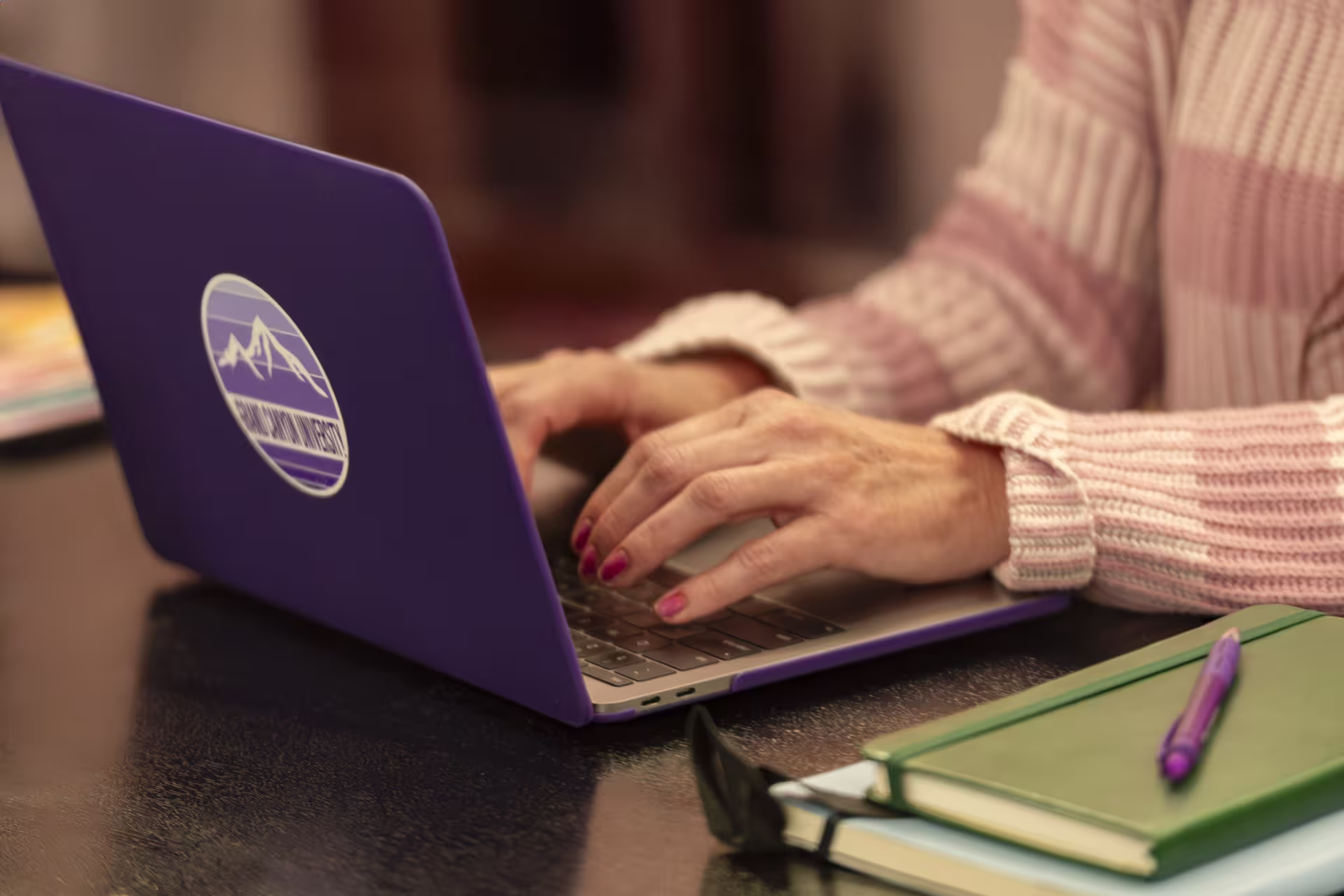 close up of hands typing on purple laptop next to notebook