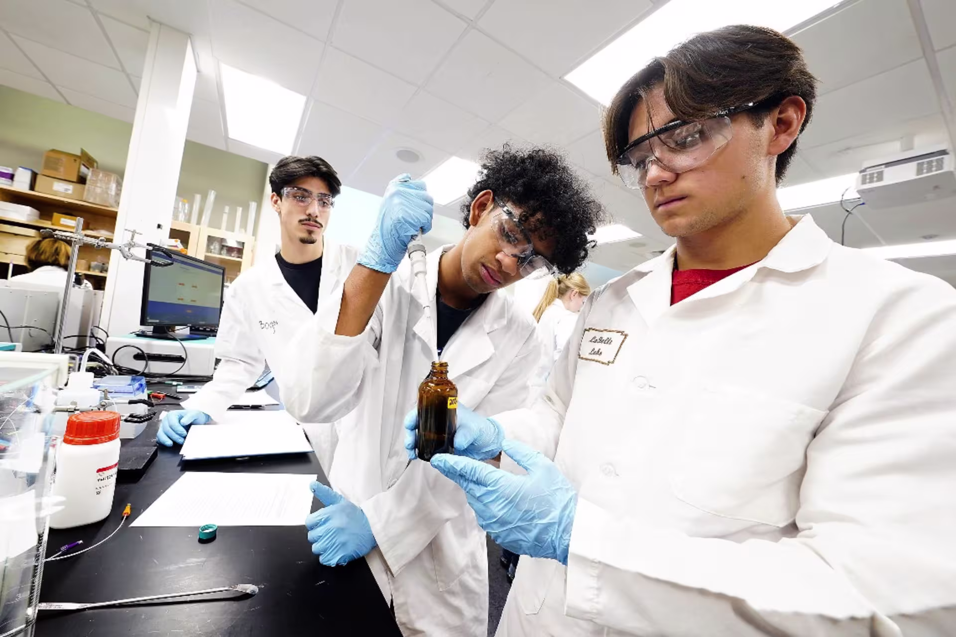 Three students in white coats and goggles working in lab setting