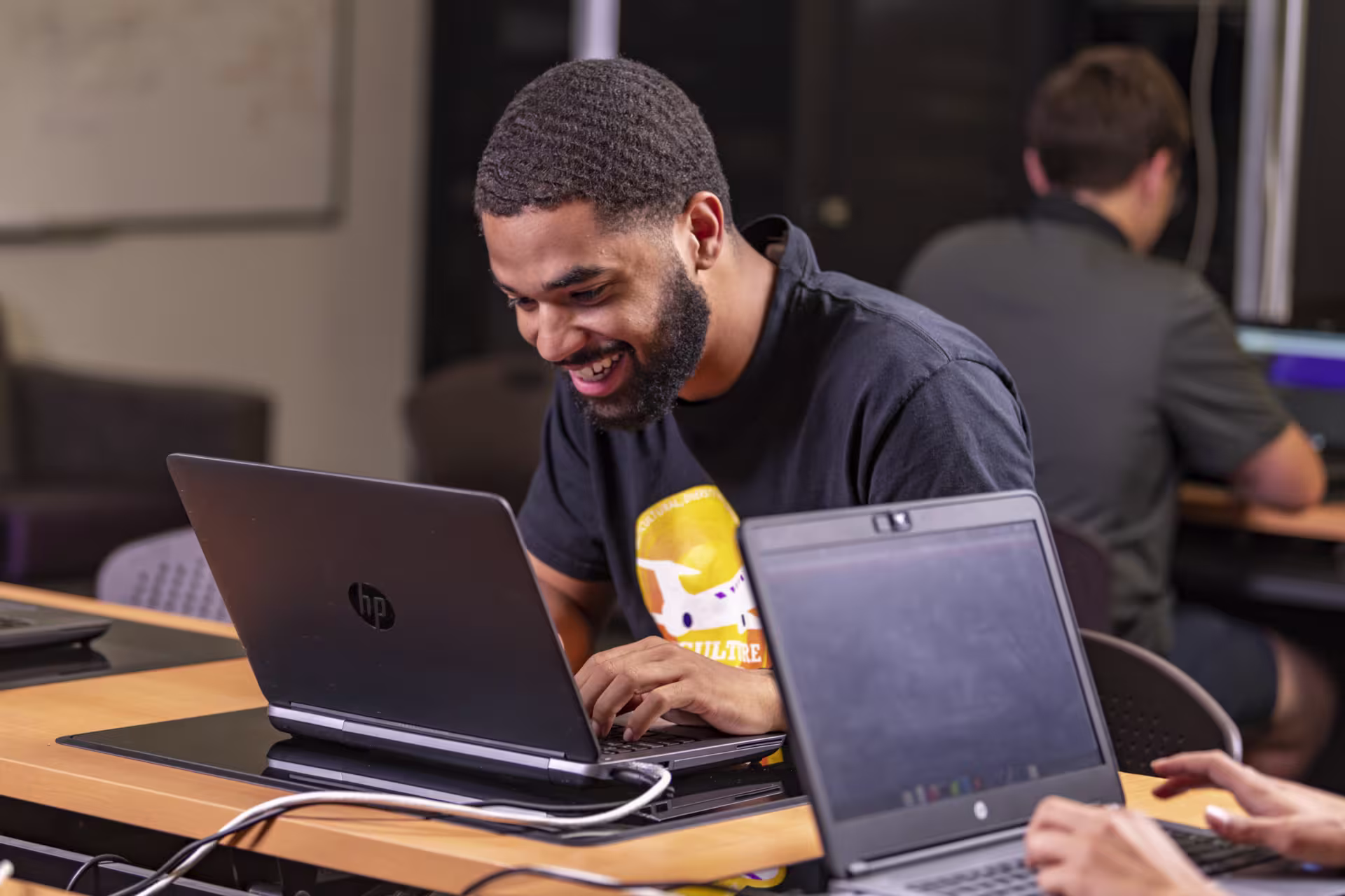 African-American male student in a black shirt smiles while looking down at laptop doing software development courses.