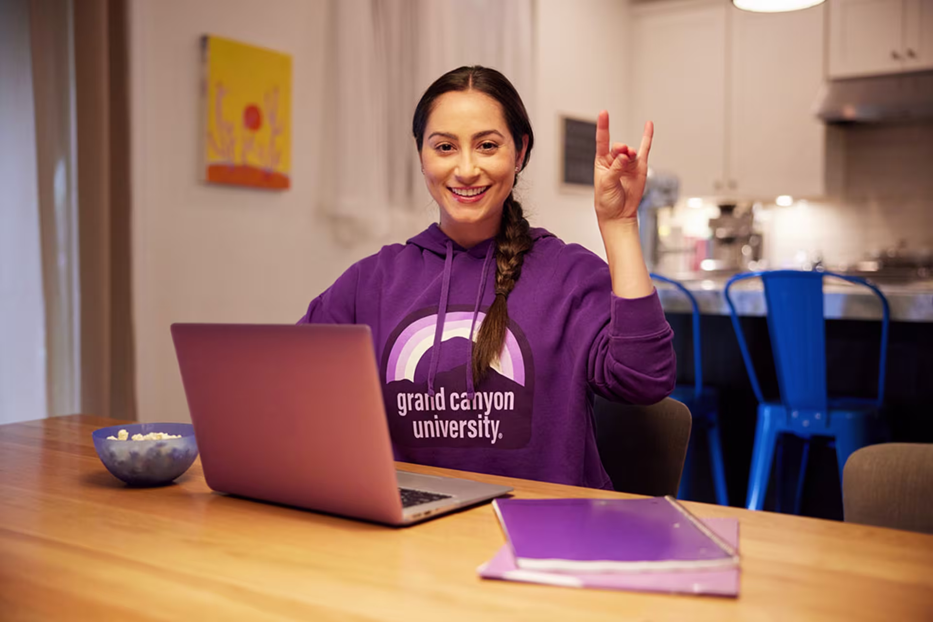 Female mental health student studying on laptop and throwing up Lopes sign in kitchen