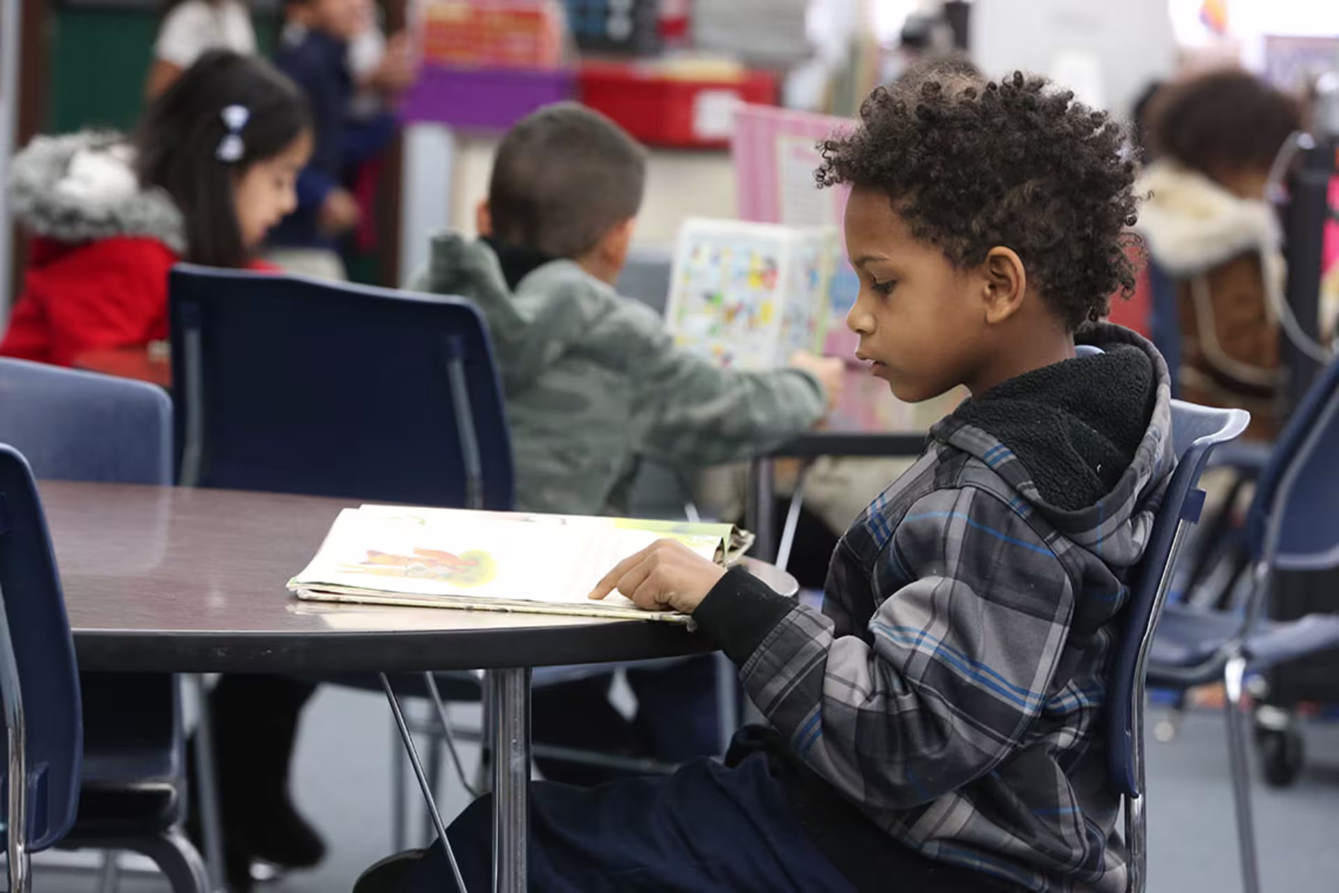 Focused shot of young male early education student reading at desk with peers around him