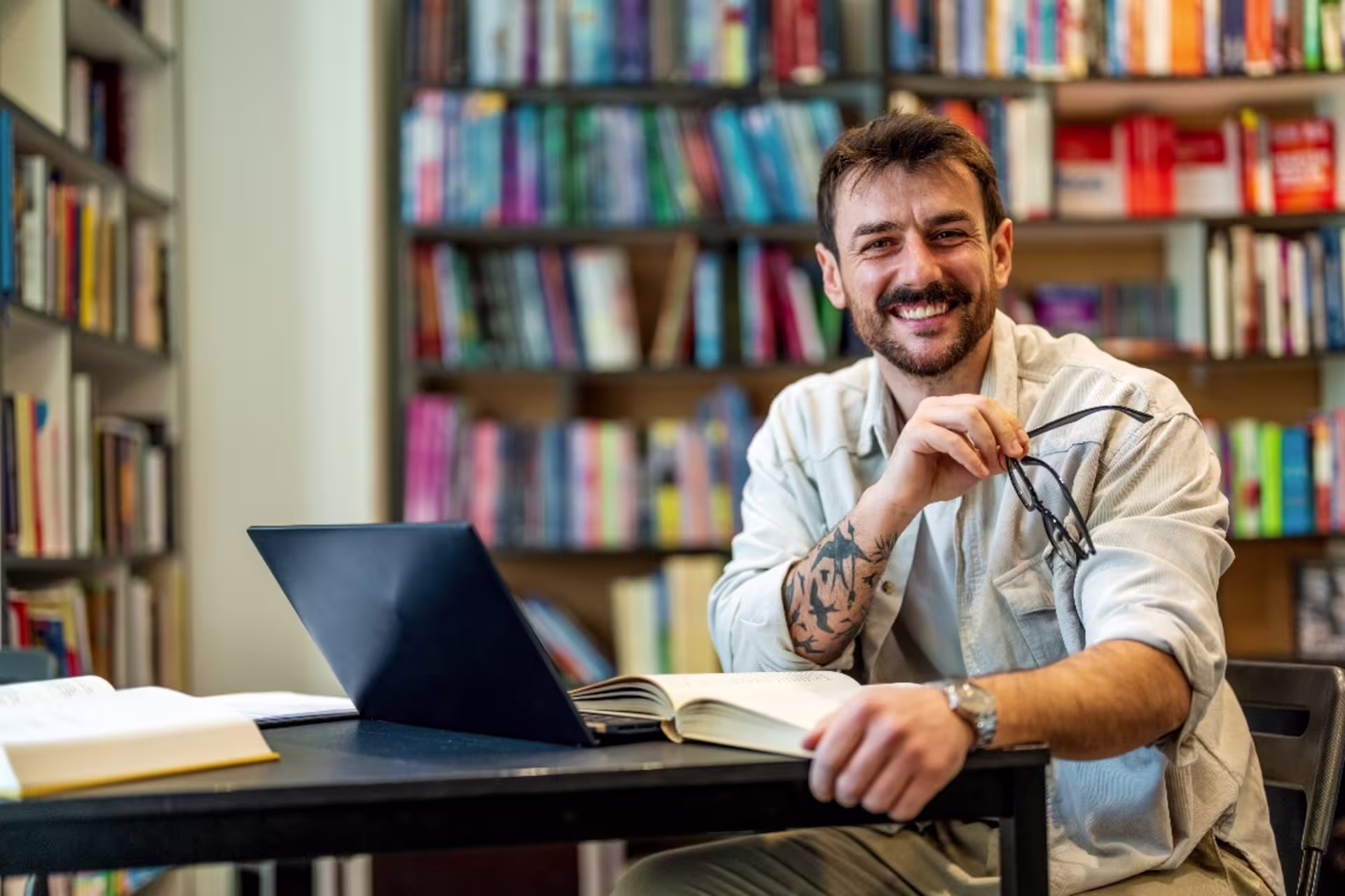 History student sitting in library with laptop while holding reading glasses