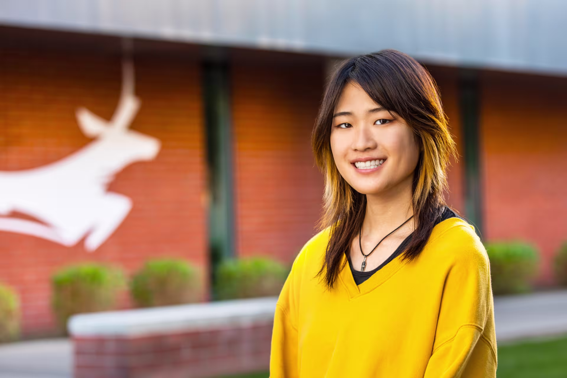 A smiling young teacher in a yellow sweater standing outside with a Grand Canyon University Lopes graphic on a brick wall in the background