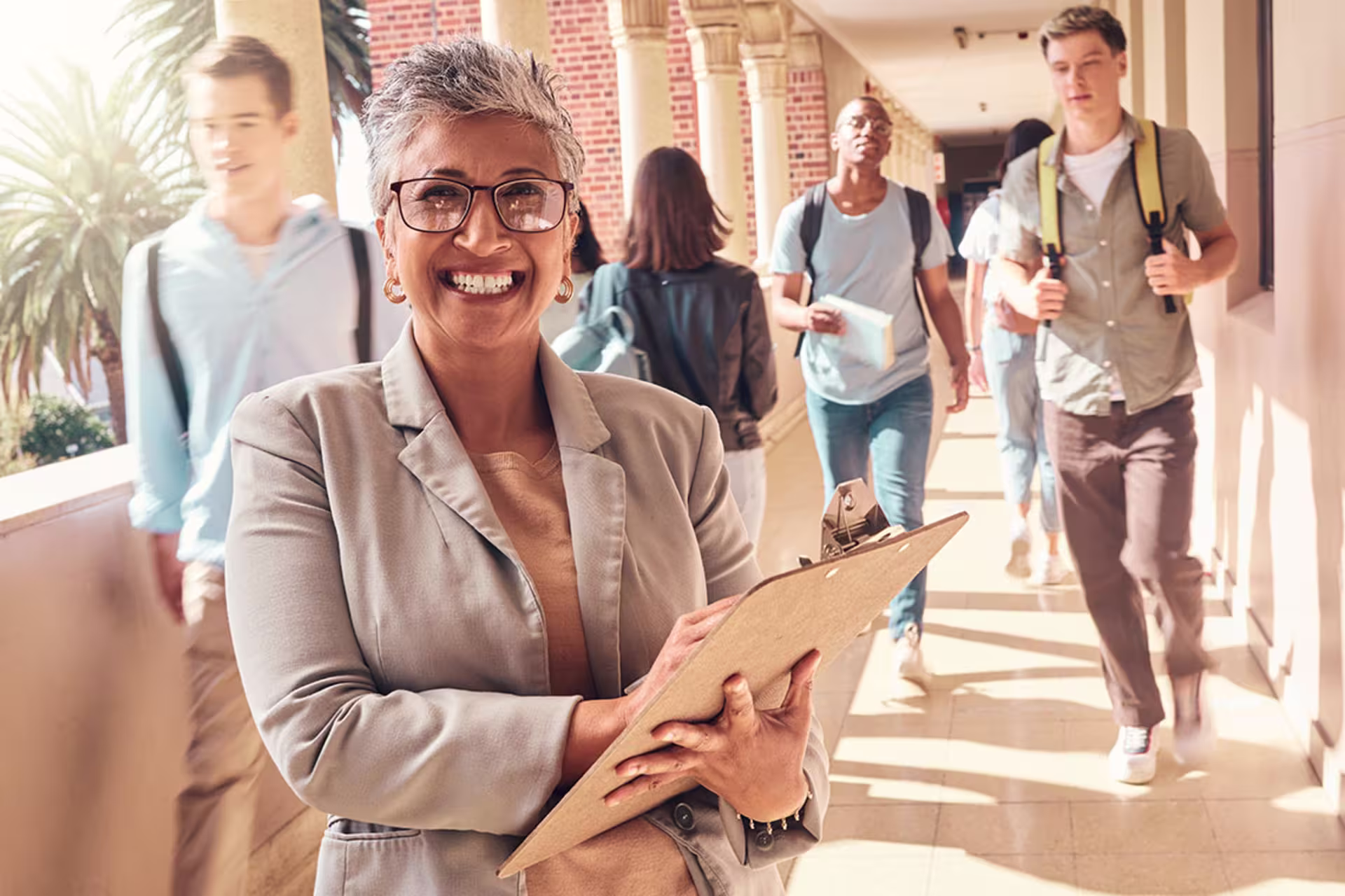 Focused shot of female academic dean walking through outside hallway while holding clipboard, smiling at camera, and students are walking through behind her