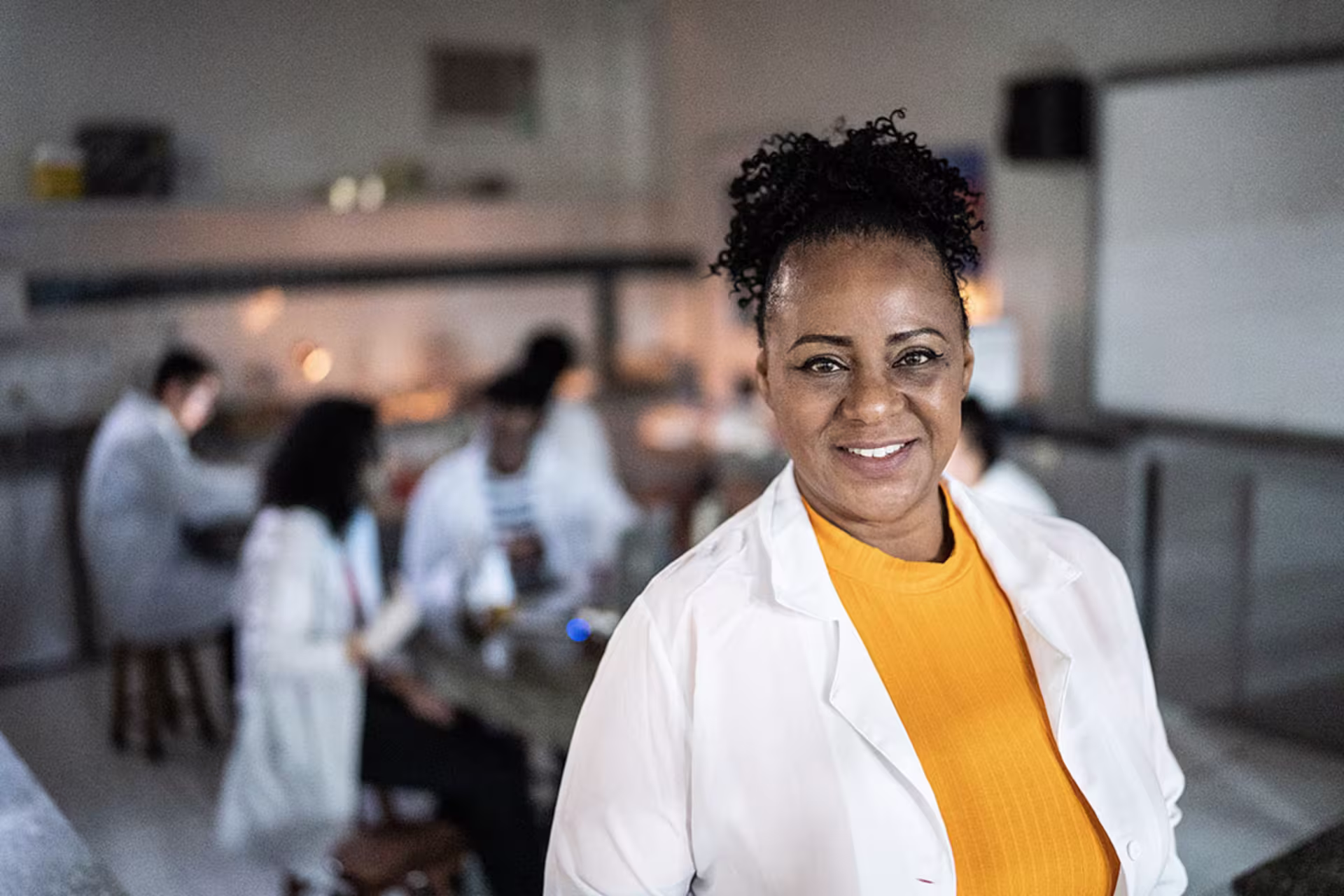 Focused shot of female biology teacher smiling at camera with classroom full of students working on labs behind her