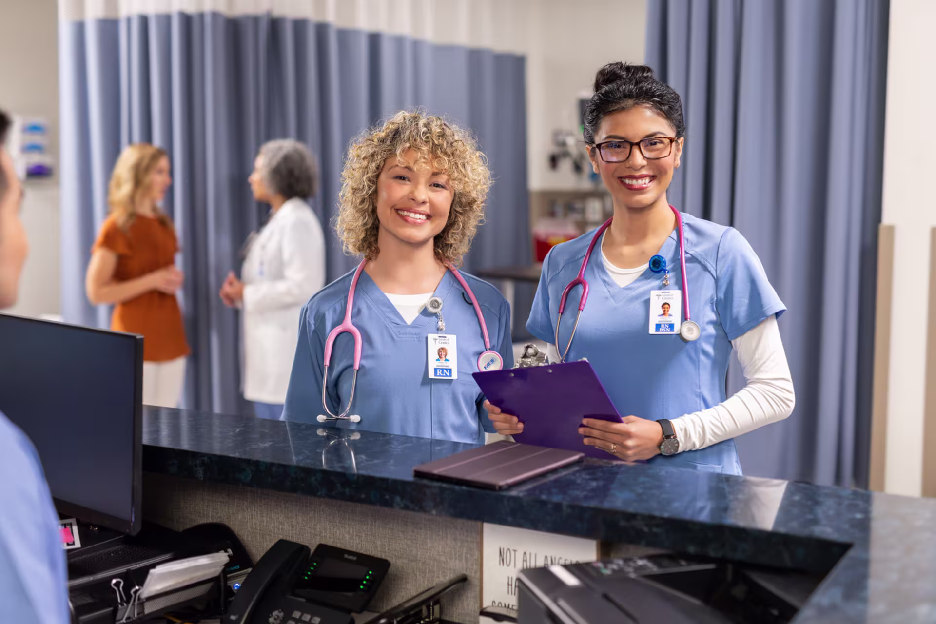 Two registered nurses in blue scrubs smiling at a hospital station, representing the Post-MSN Nursing Certificate program.