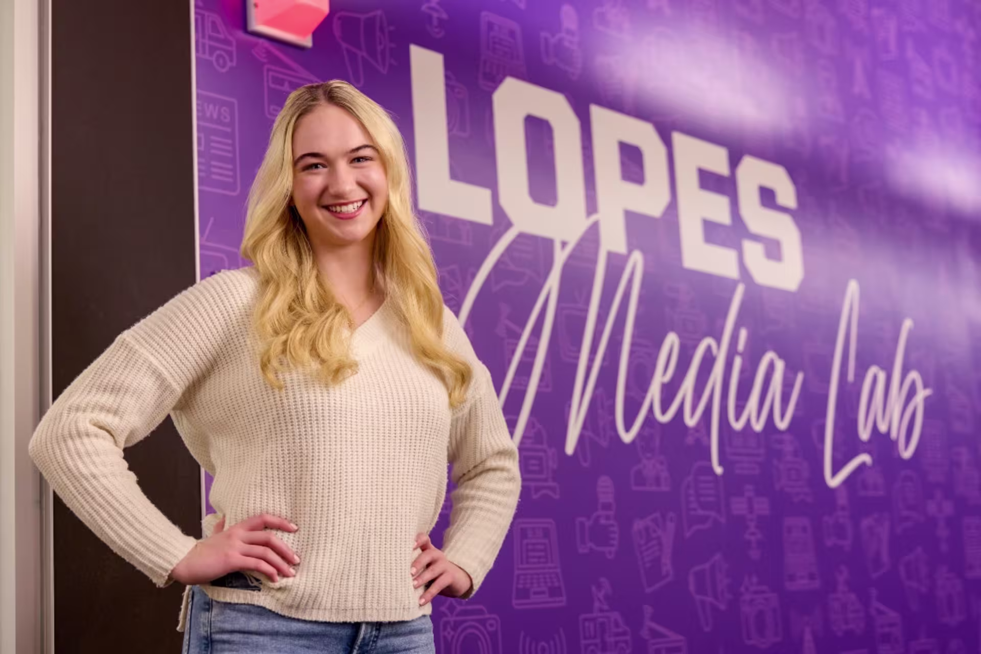 Student in white sweater standing in front of the Lopes Media Lab