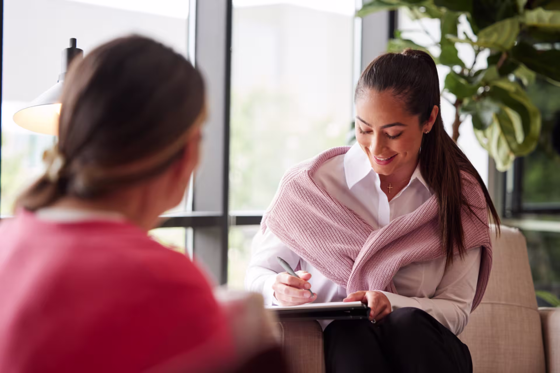 Counselor in a pink sweater sitting with a client and taking notes