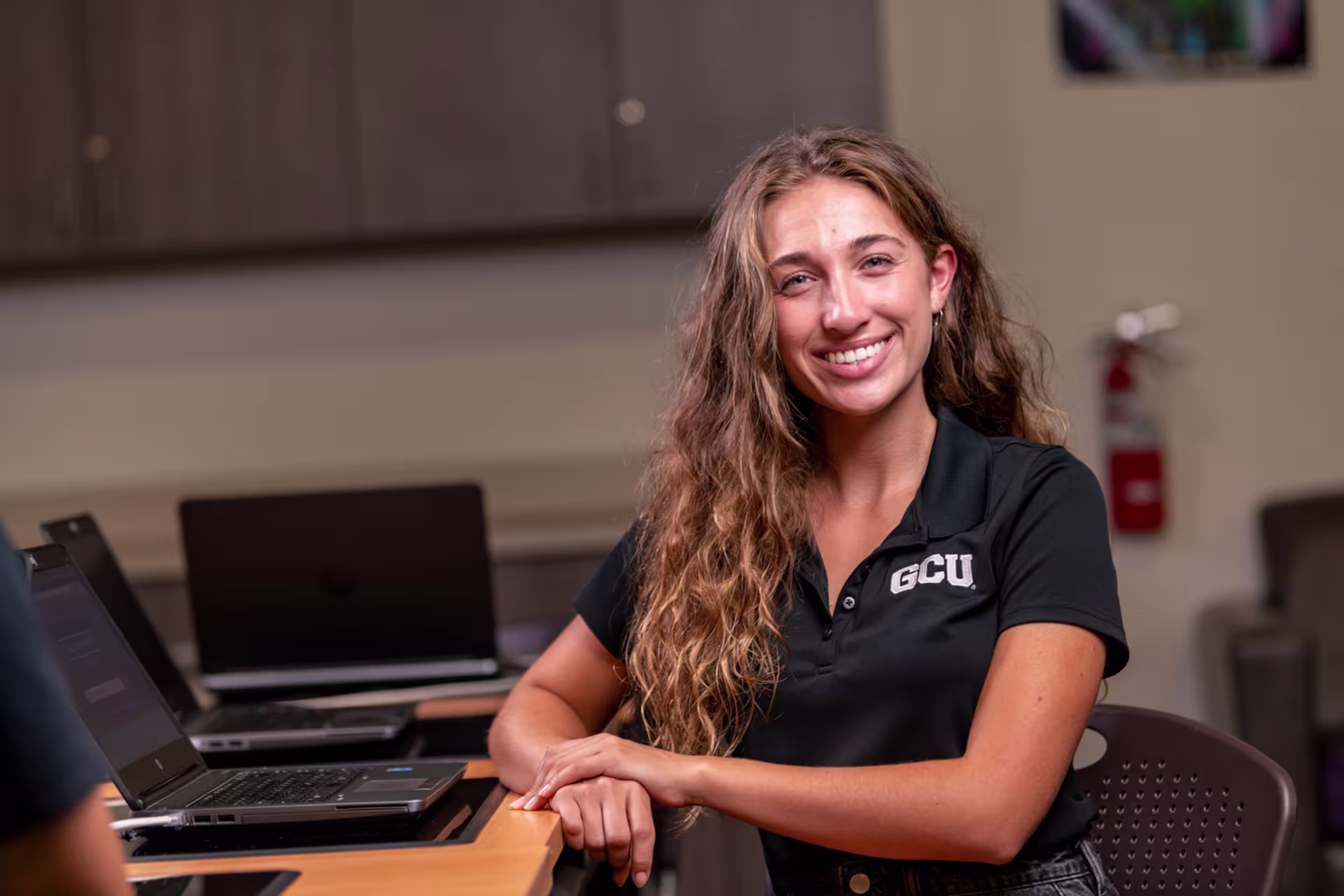 Female science student in GCU shirt smiling at desk in classroom