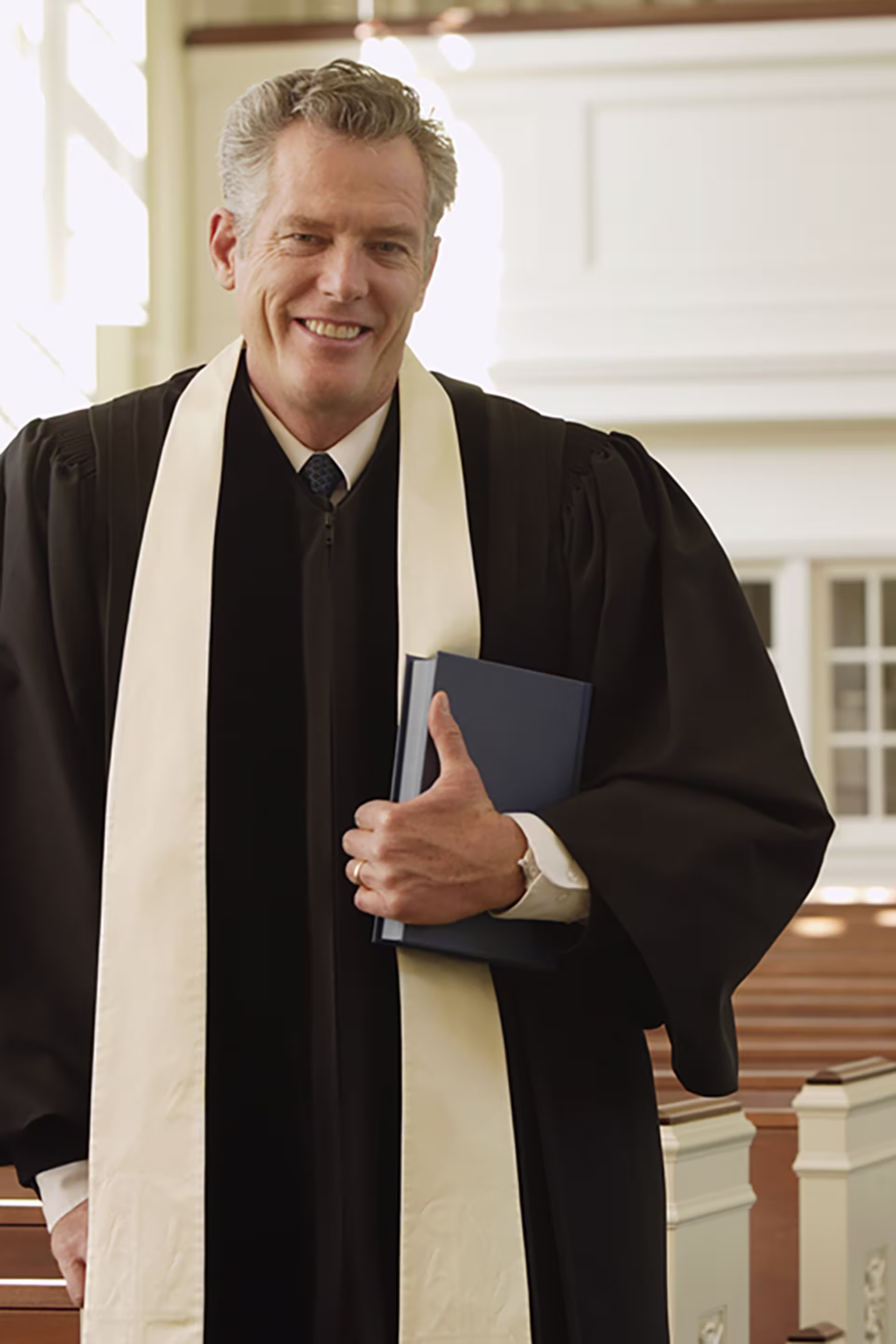 older male preacher holding bible in well-lit church of empty pews