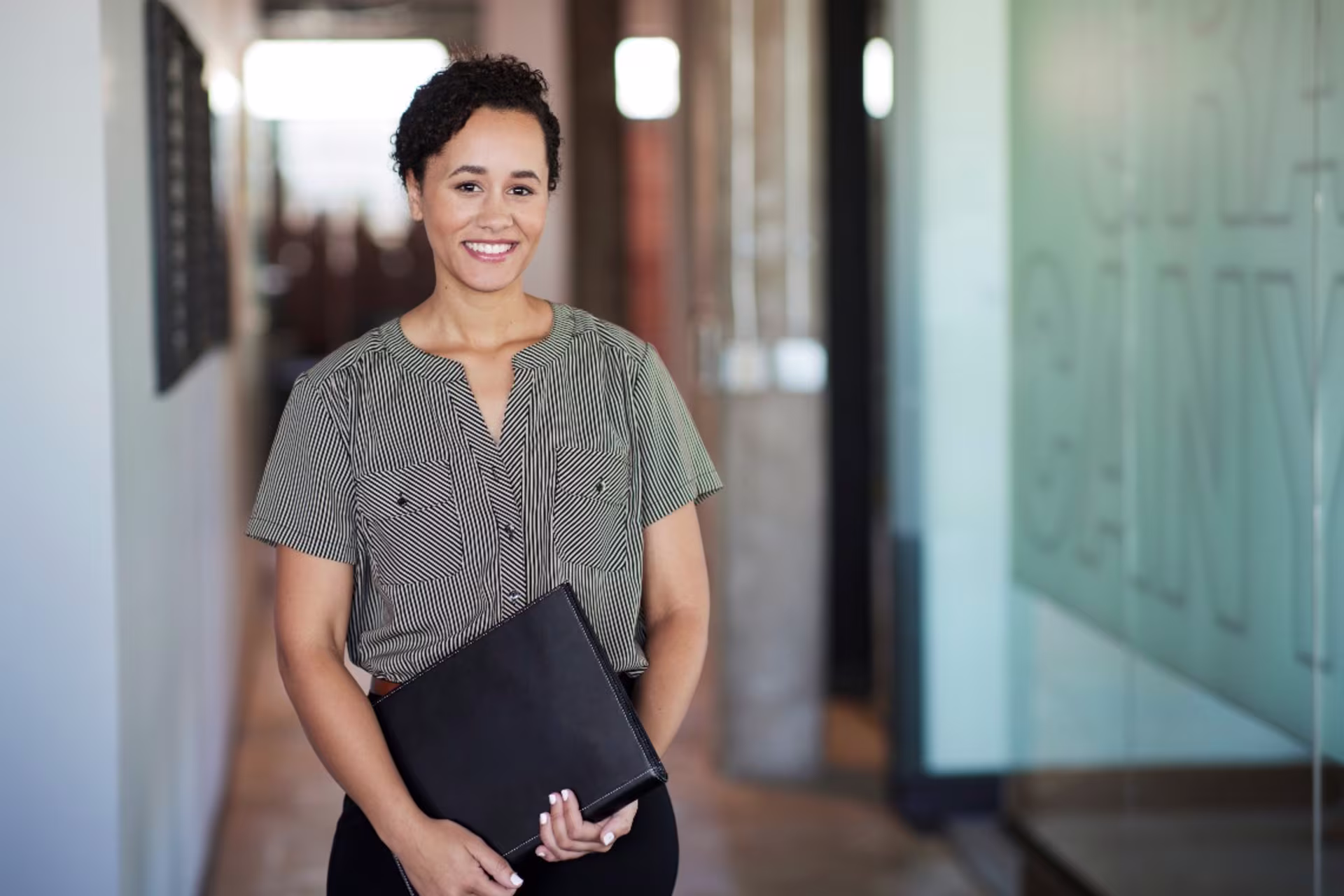 short curly-haired female bachelor-level accounting graduate interns in a firm while holding a folder in a hallway