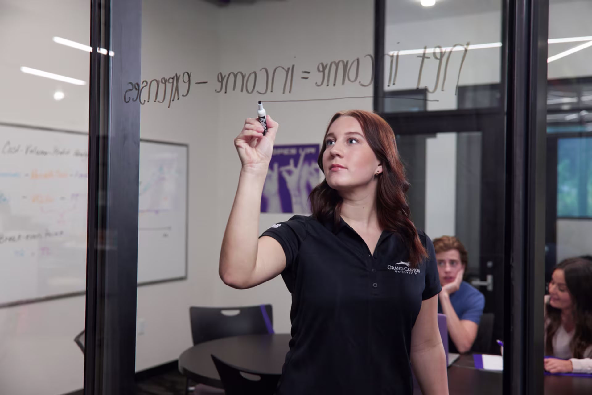 redhead female accounting student in a black GCU branded shirt writing net income equations on a board