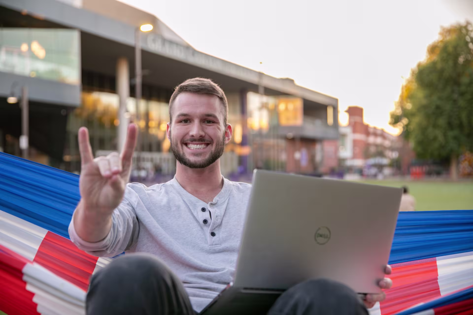 man sitting in patriotic hammock smiles while holding lopes up sign with open laptop on lap