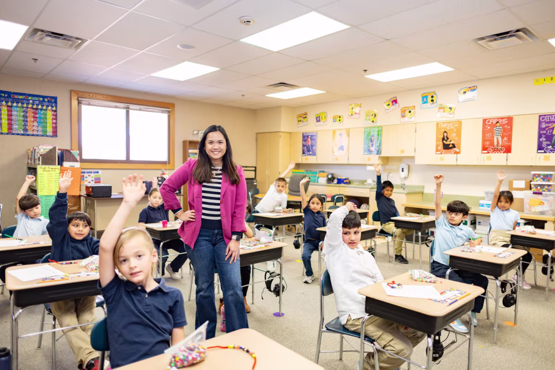 Young female school teacher standing in elementary education classroom of students with hands raised at their desks