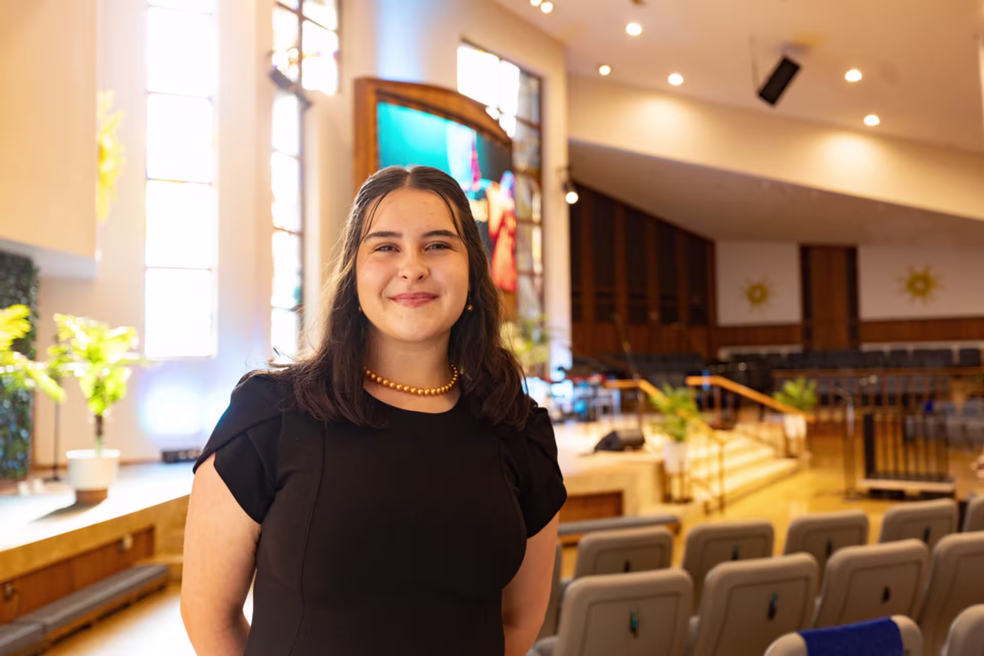 Brunette white Christian theology graduate certificate student in a black dress stands in an empty church filled with bright light