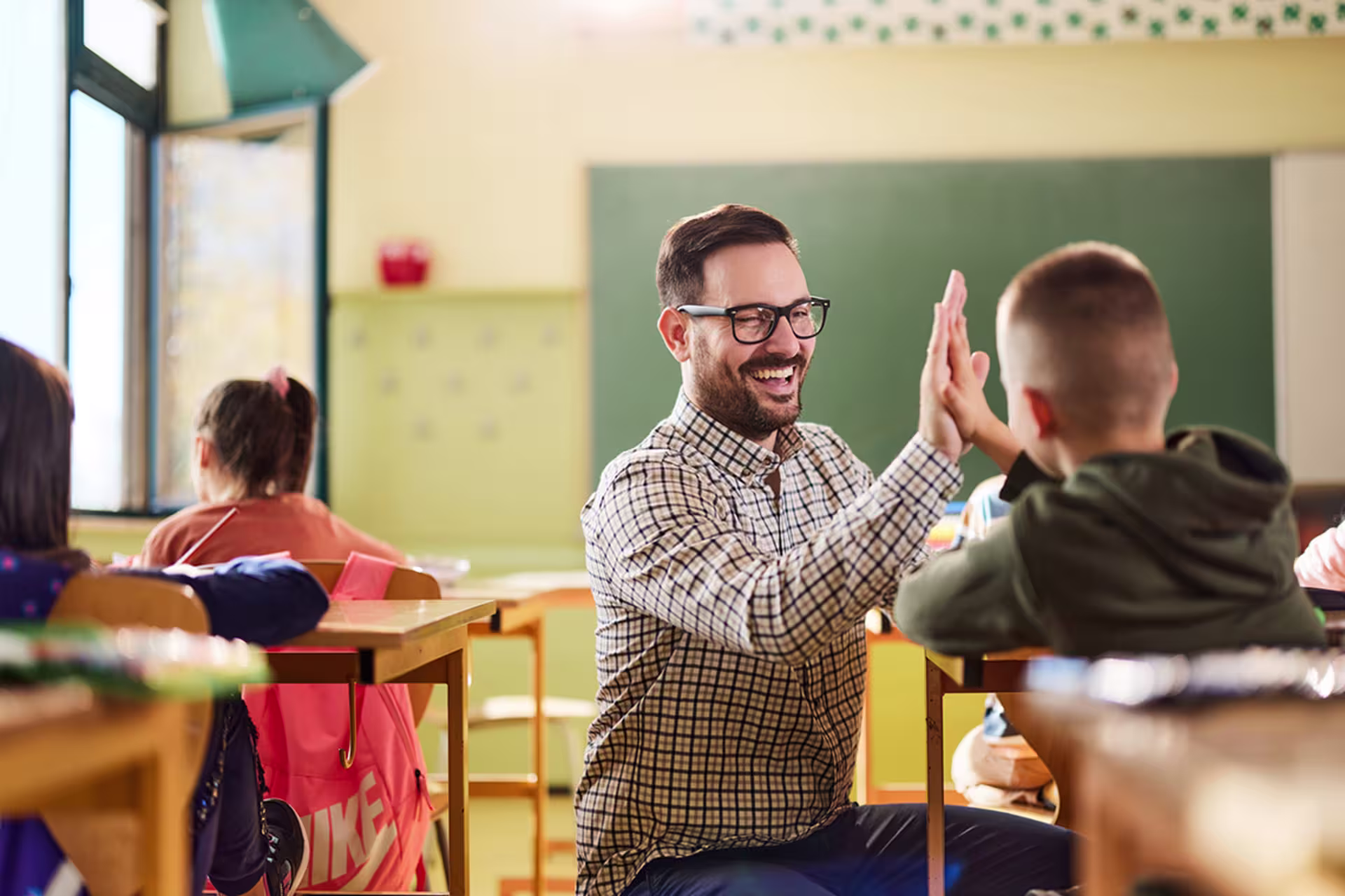 bearded white male elementary education teacher in black glasses gives a student a high five while crouching at their desk