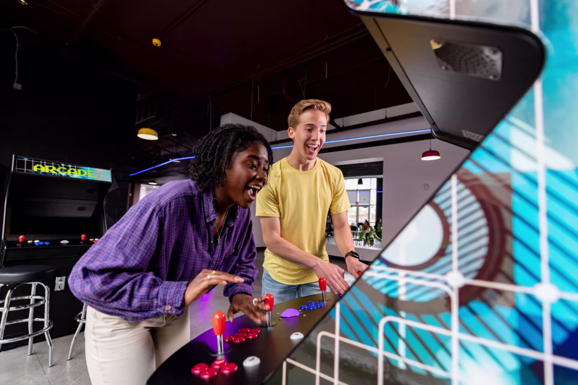 Enthusiastic African-American college student plays arcade game in the college recreation space with a blonde white guy watching in excitement 