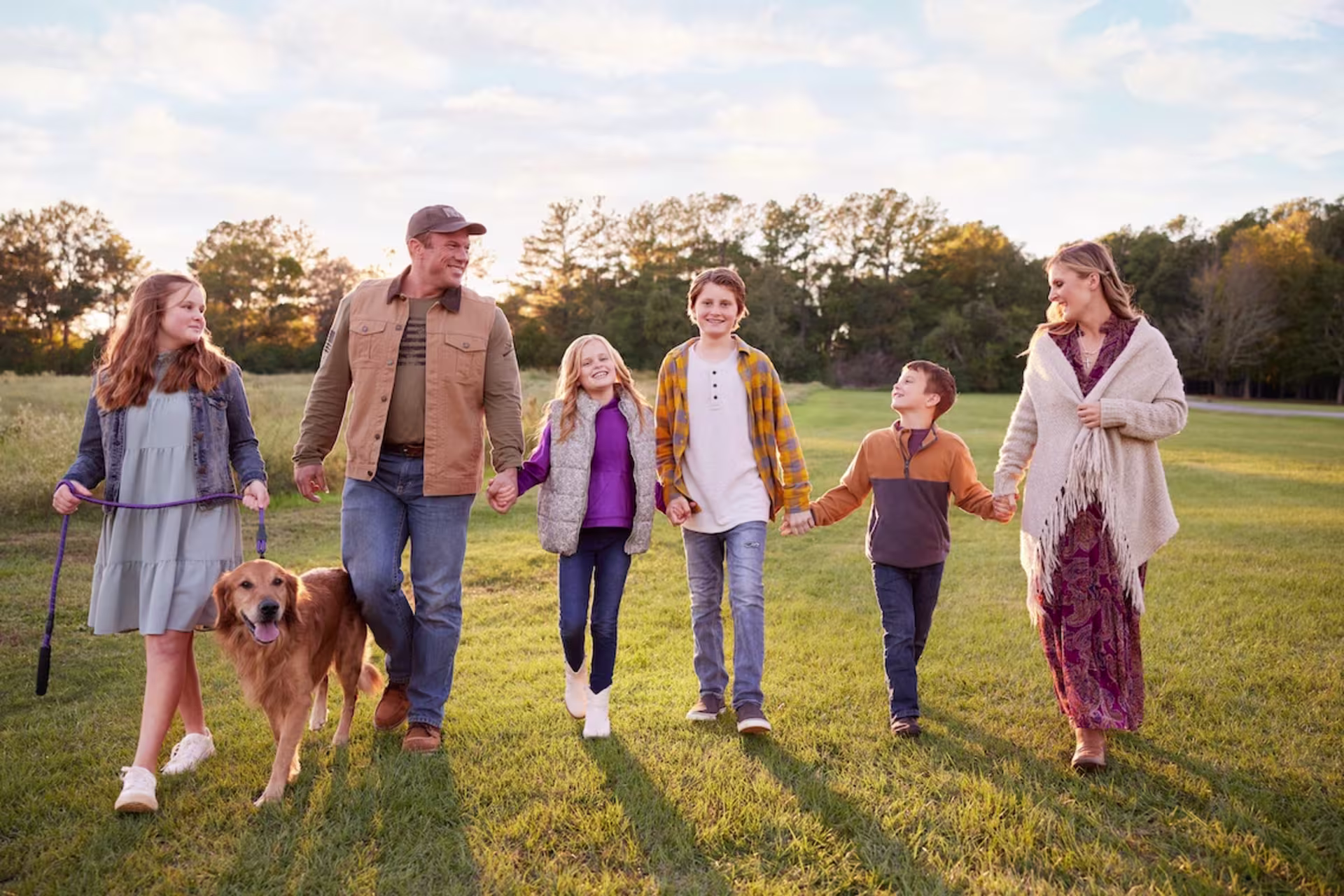 family with dog smiling while holding hands walking across field.