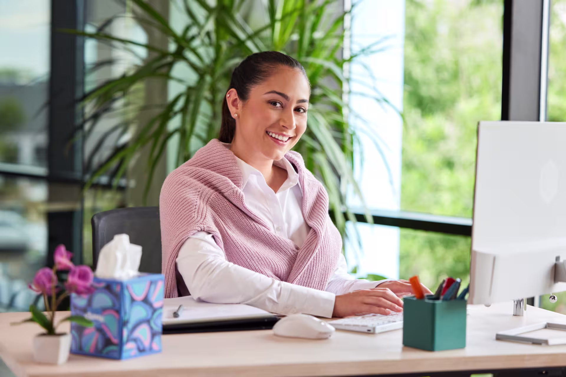 Social worker in pink sweater sitting and smiling at a desk while working on a laptop.