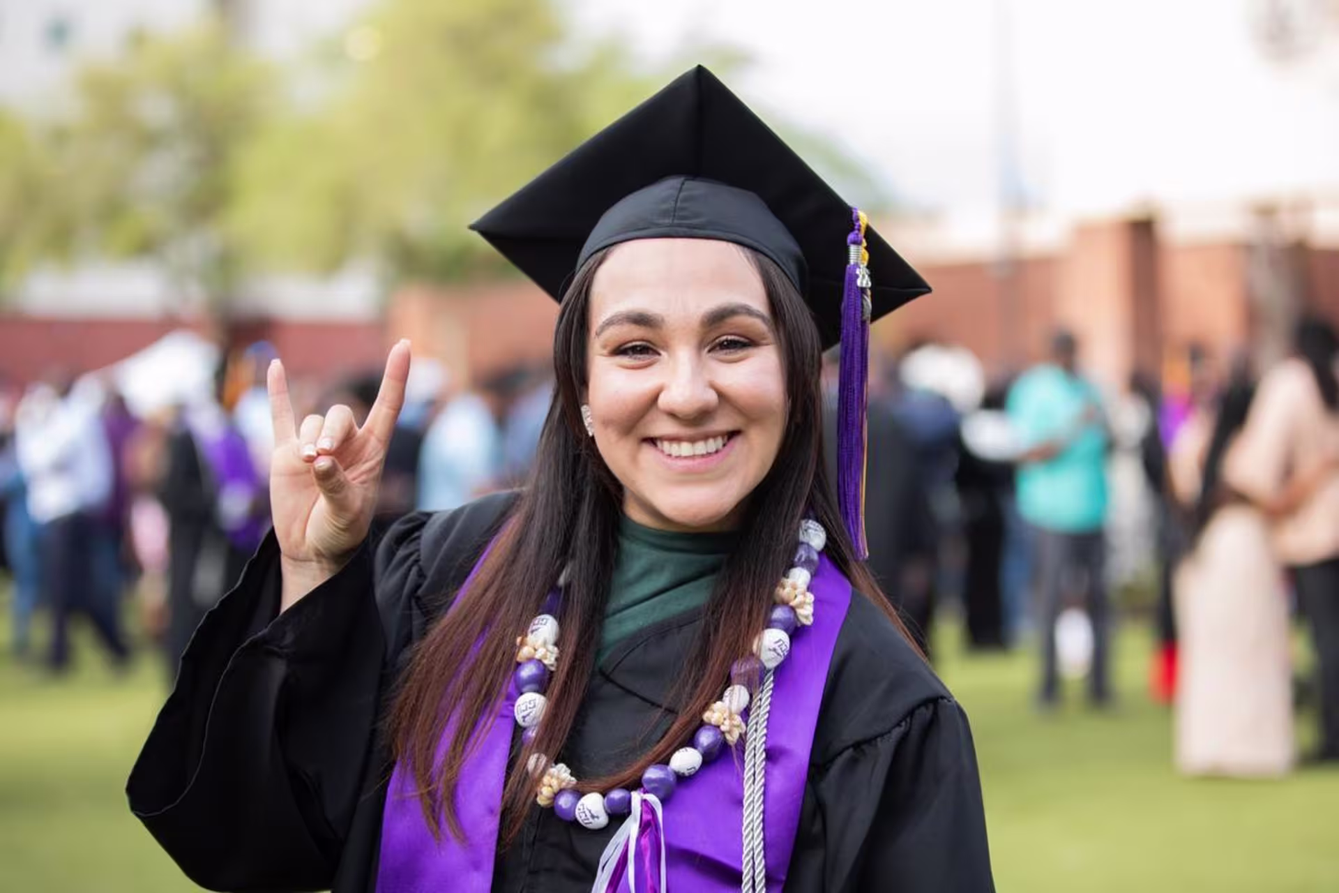 Female GCU graduate in cap and gown smiling at the camera outdoors, holding up the Lopes hand sign.