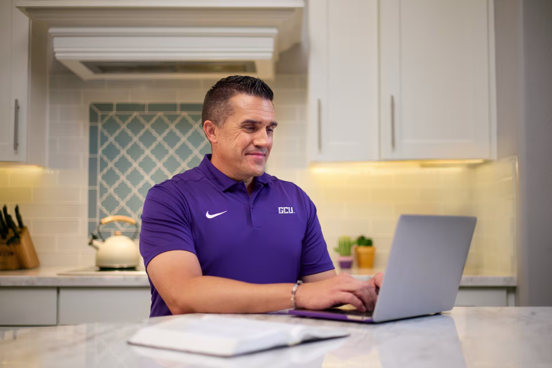 Male graduate artificial intelligence certificate student in a purple polo working from home on a laptop.
