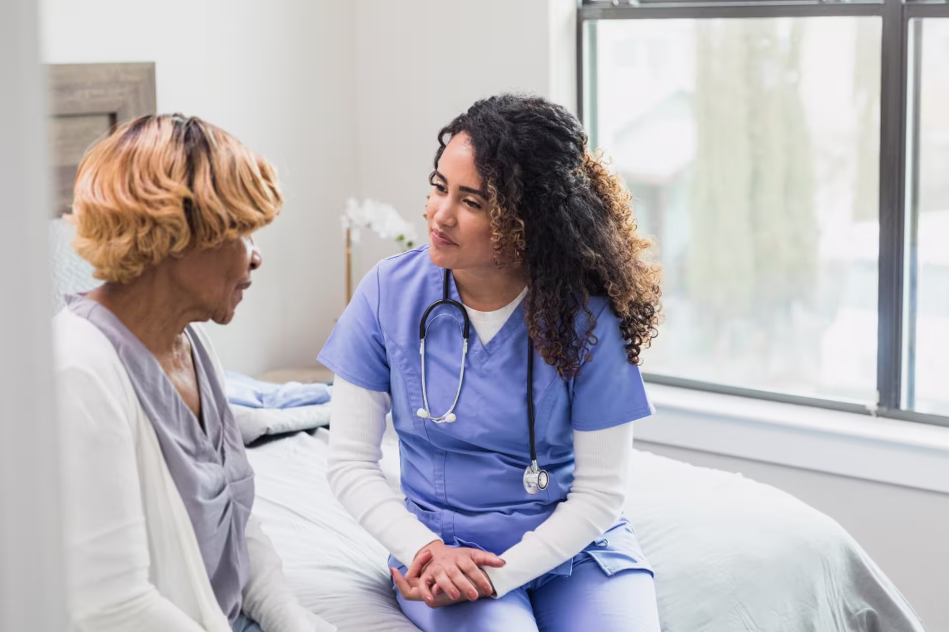 Psychiatric mental health nurse practitioner sitting on a bed while talking with an older woman.