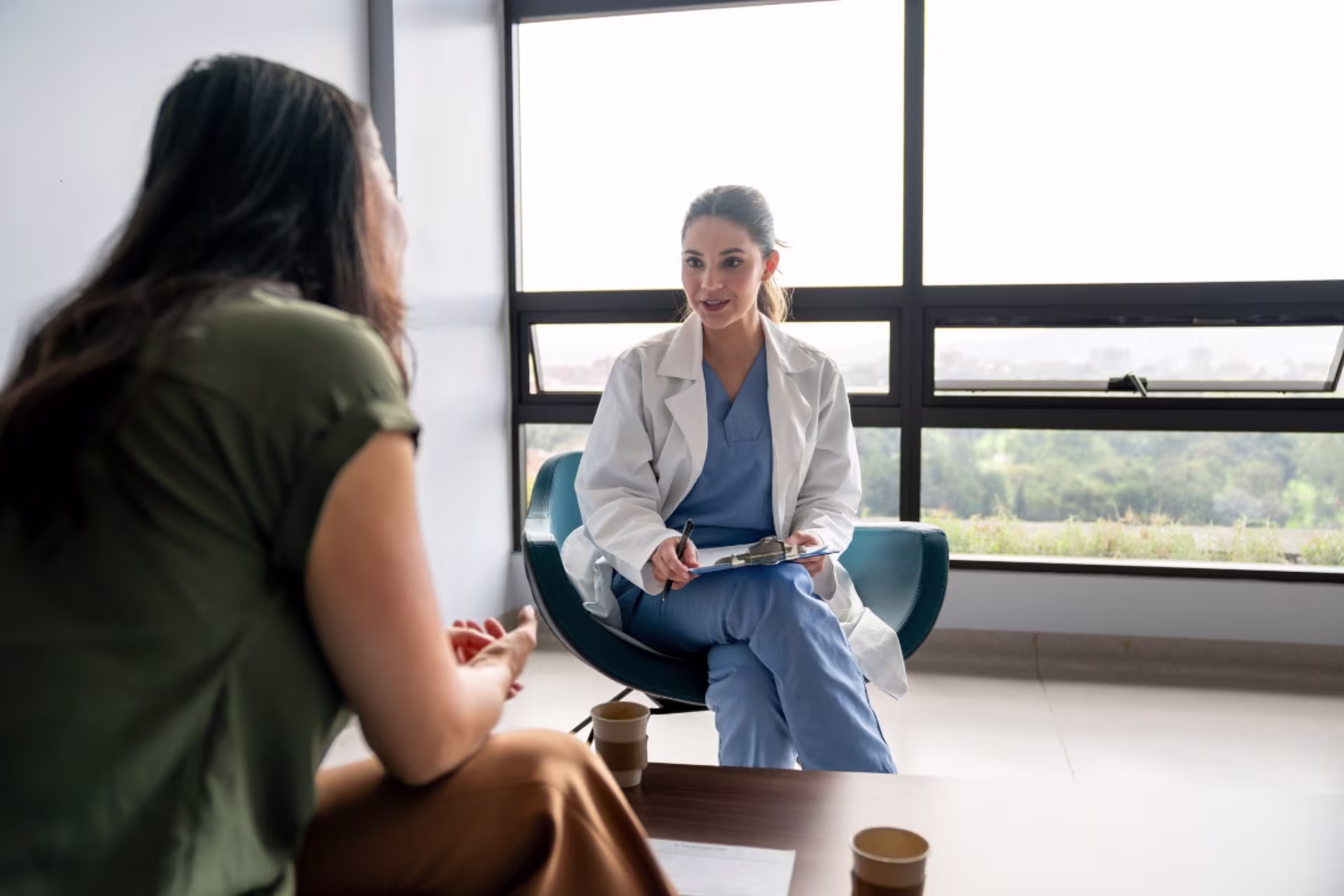 PMHNP certificate holder in light blue scrubs taking notes while talking with a patient.