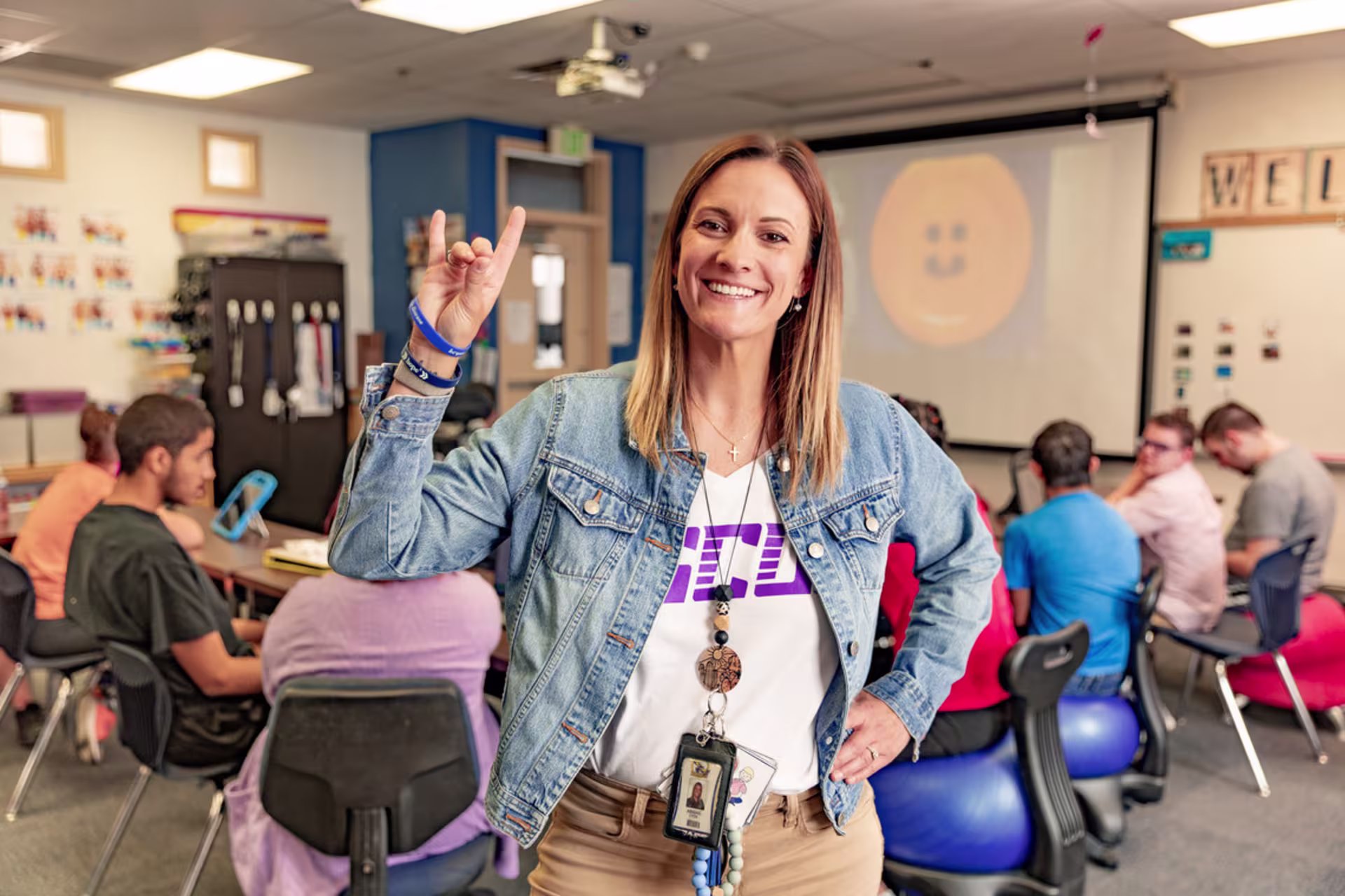 Female secondary education teacher holds up the Lopes hand signal in a classroom with high school students.