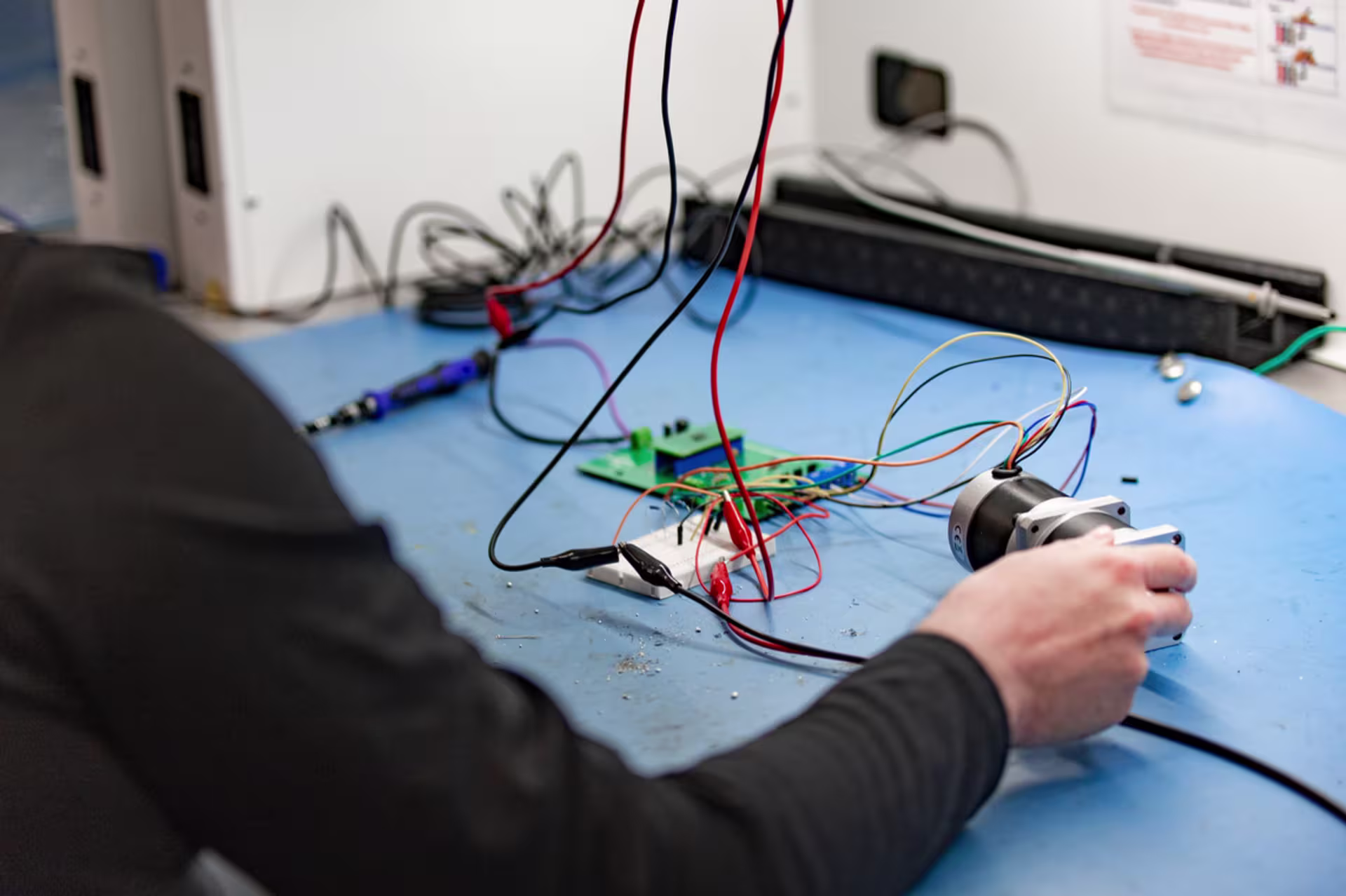 Person working at an electronics workstation, holding a component connected to a circuit board with multiple wires and tools on a blue work surface.