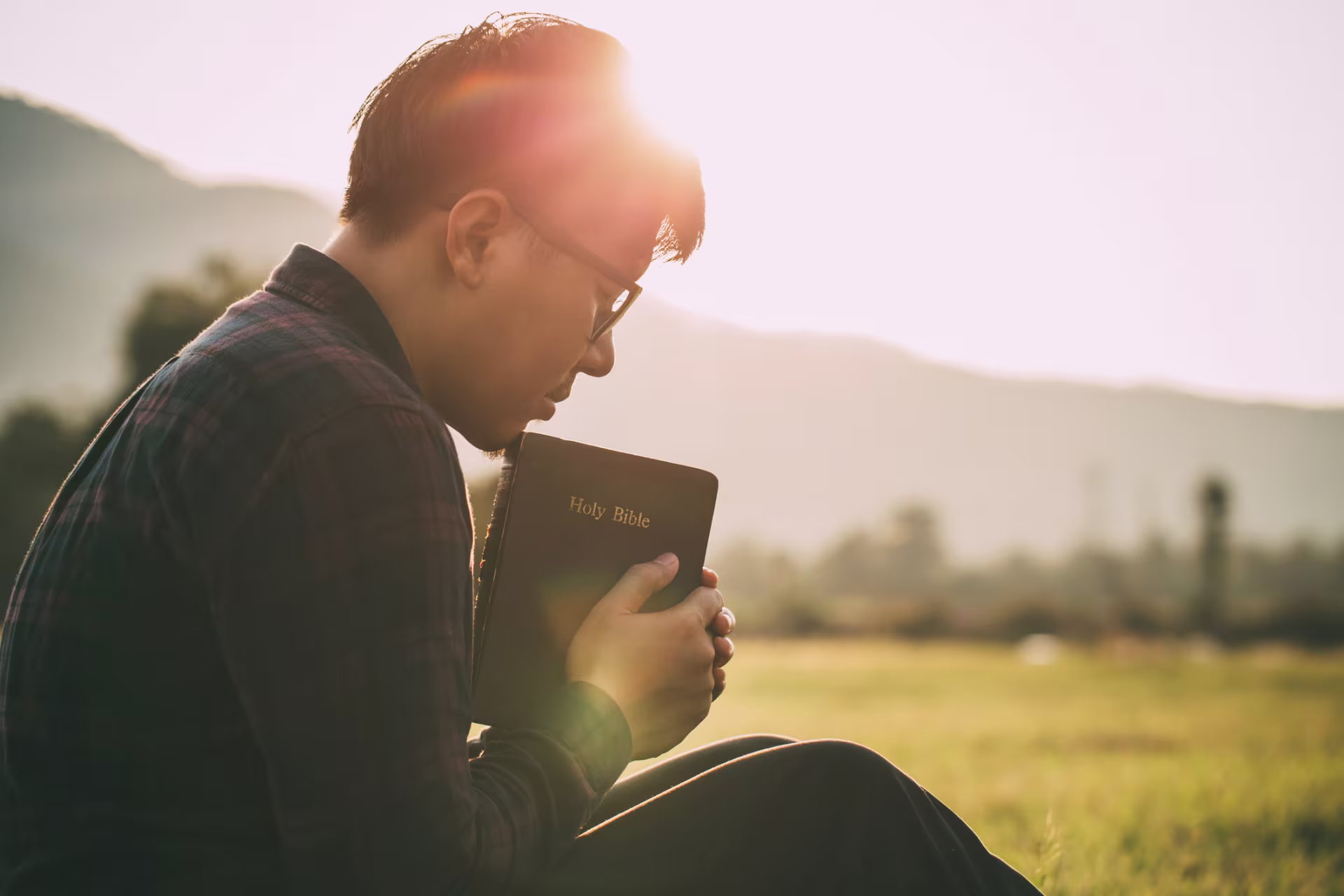 Person sitting outdoors at sunset, holding a Holy Bible close while reflecting on Jesus' life in a peaceful grassy field.