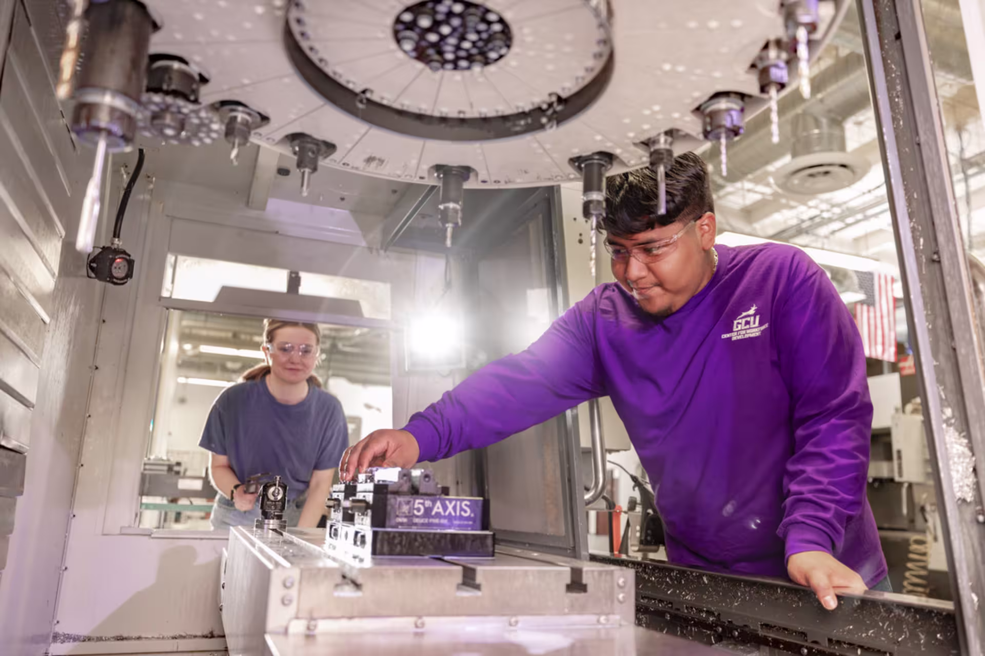 Male trade certificate student adjusts tool settings inside a 5-axis cnc machine, learning how to operate a machine, hands-on.