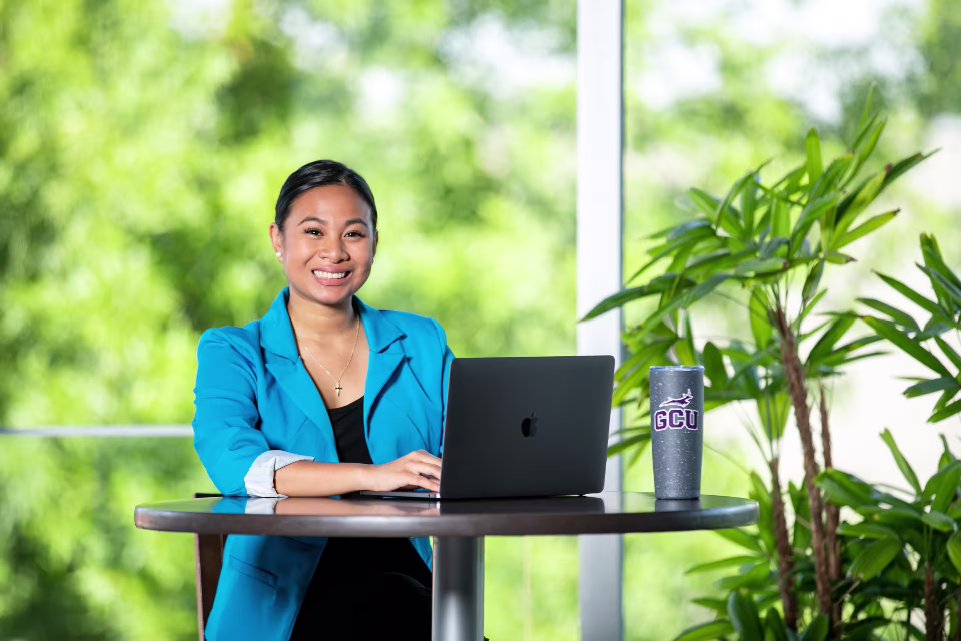 A woman in professional blue blazer attire with hair sleeked back smiles while typing on black laptop for her PhD in educational technology online course.