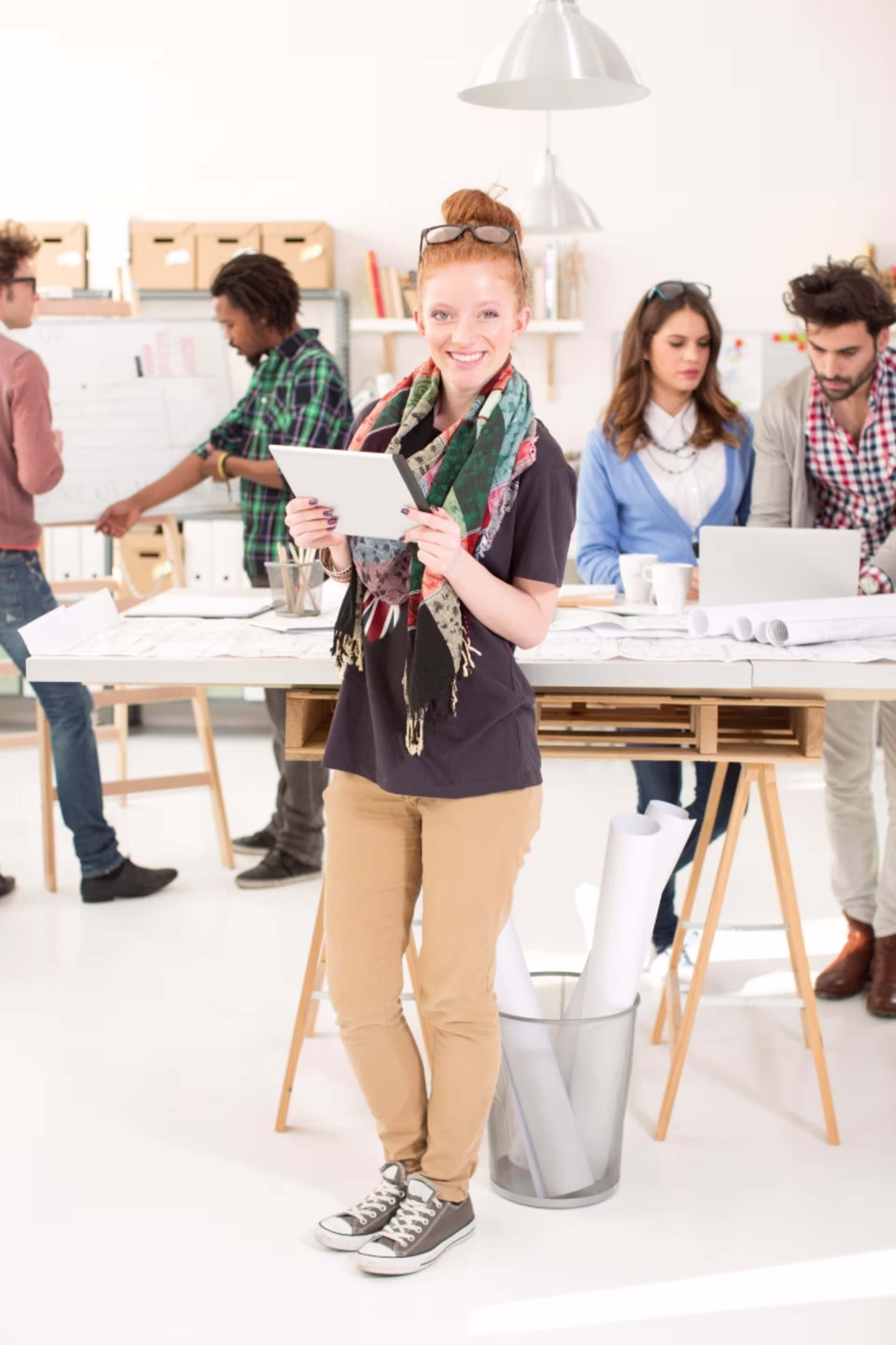 A female stands with a tablet in front of communications colleagues working together at desks.