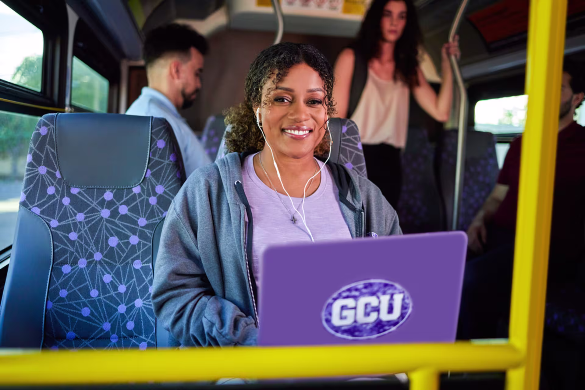 Smiling woman on a bus is taking an online class for her GCU degree on her laptop, wearing earphones and a hoodie.