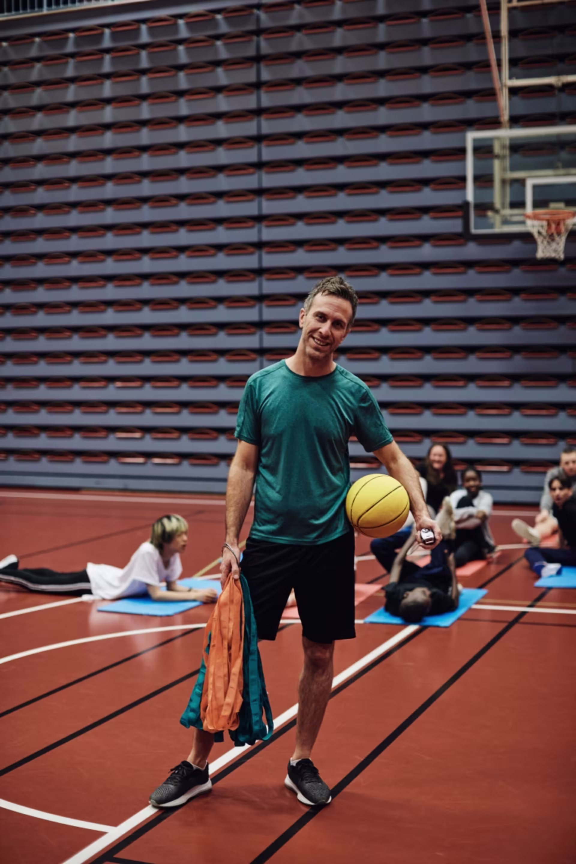 Male teacher standing in a gym holding a basketball with students seated in the background.