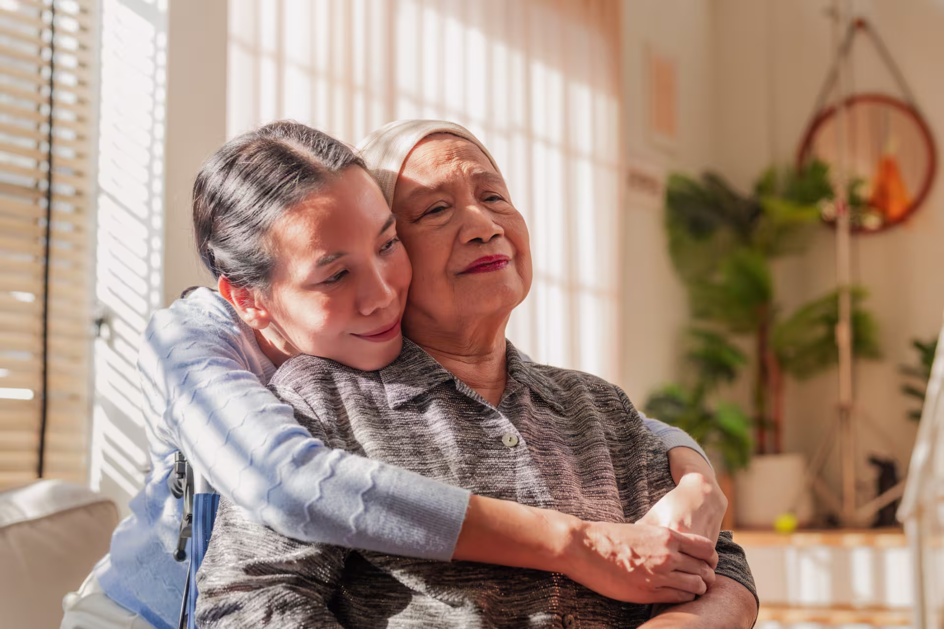 A person is sitting in a sunlit living room while another person wraps their arms around them from behind in an affectionate gesture.