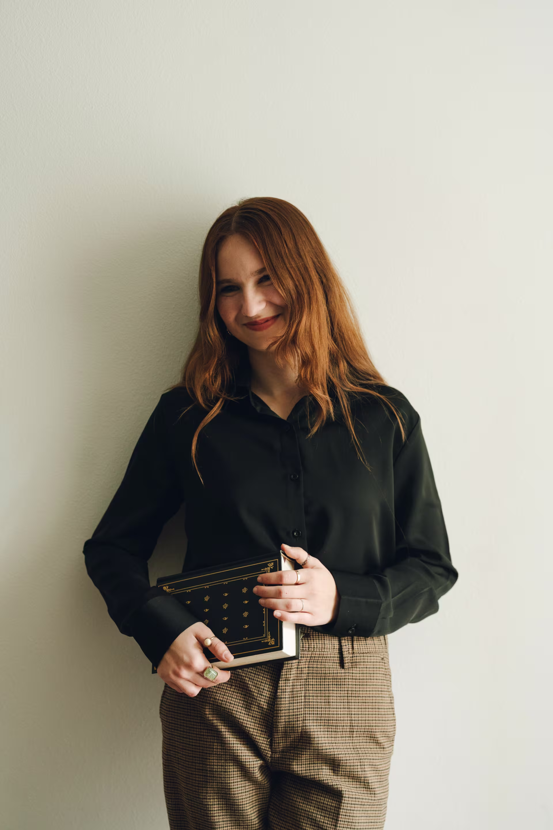 A person wearing a dark long-sleeve shirt and brown checkered pants is holding a closed book with a decorative cover against a plain light-colored wall.