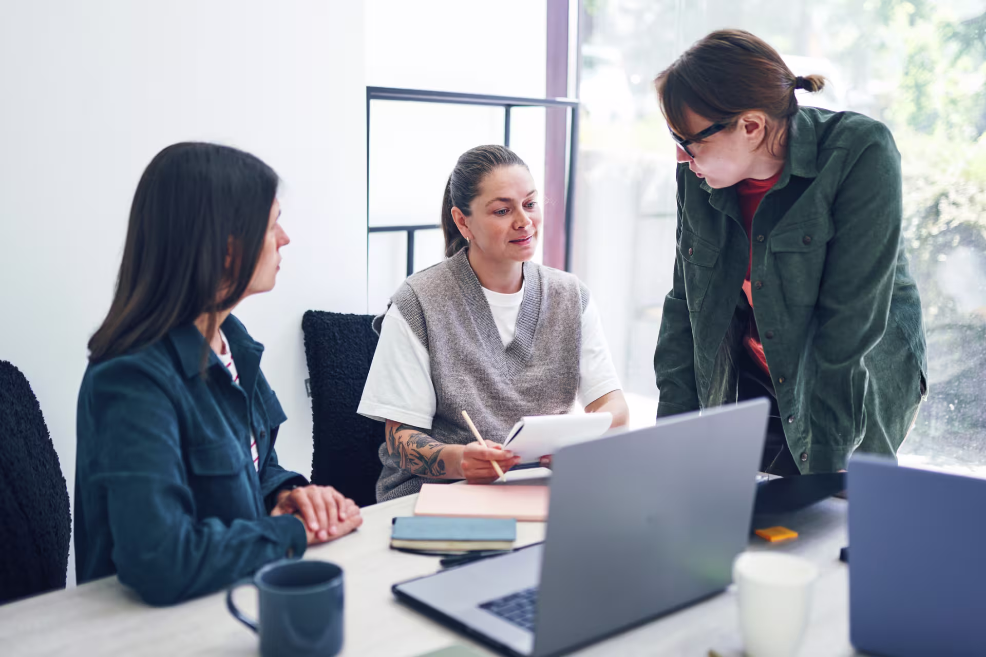 Three people sit together at a table with laptops and notebooks, engaged in a discussion.