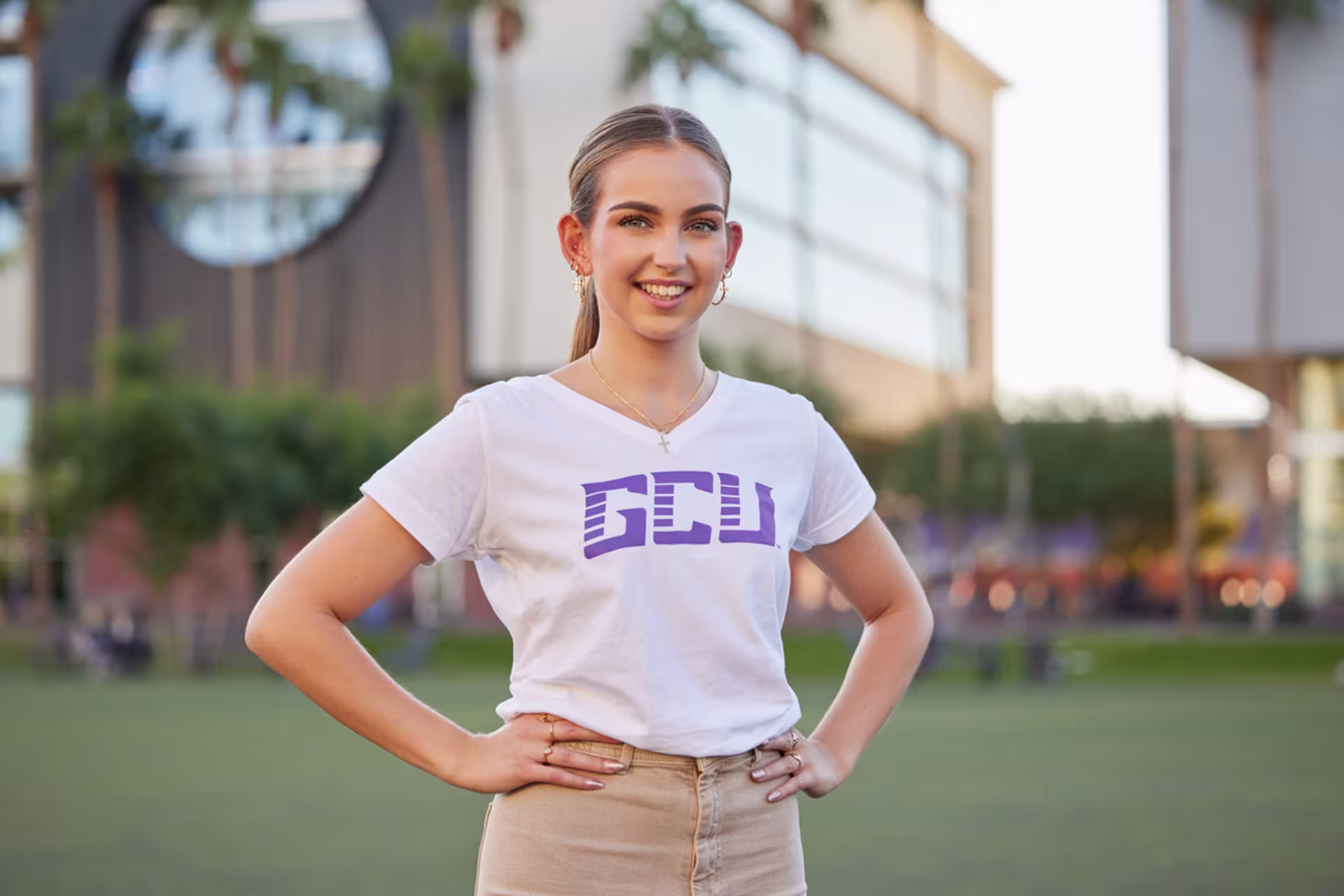 Student in white GCU shirt standing on campus with her arms on her hips and smiling