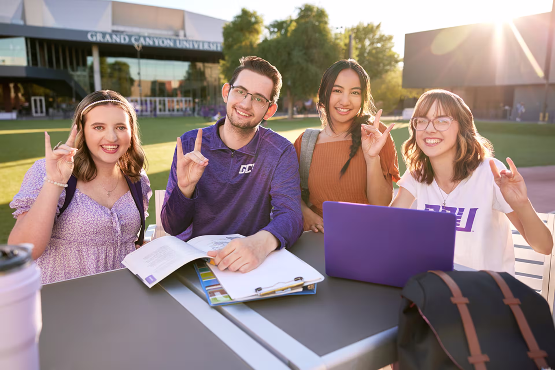 Four GCU students sitting outdoors on campus lawn, studying with laptops and notebooks in front of campus buildings on a sunny day.