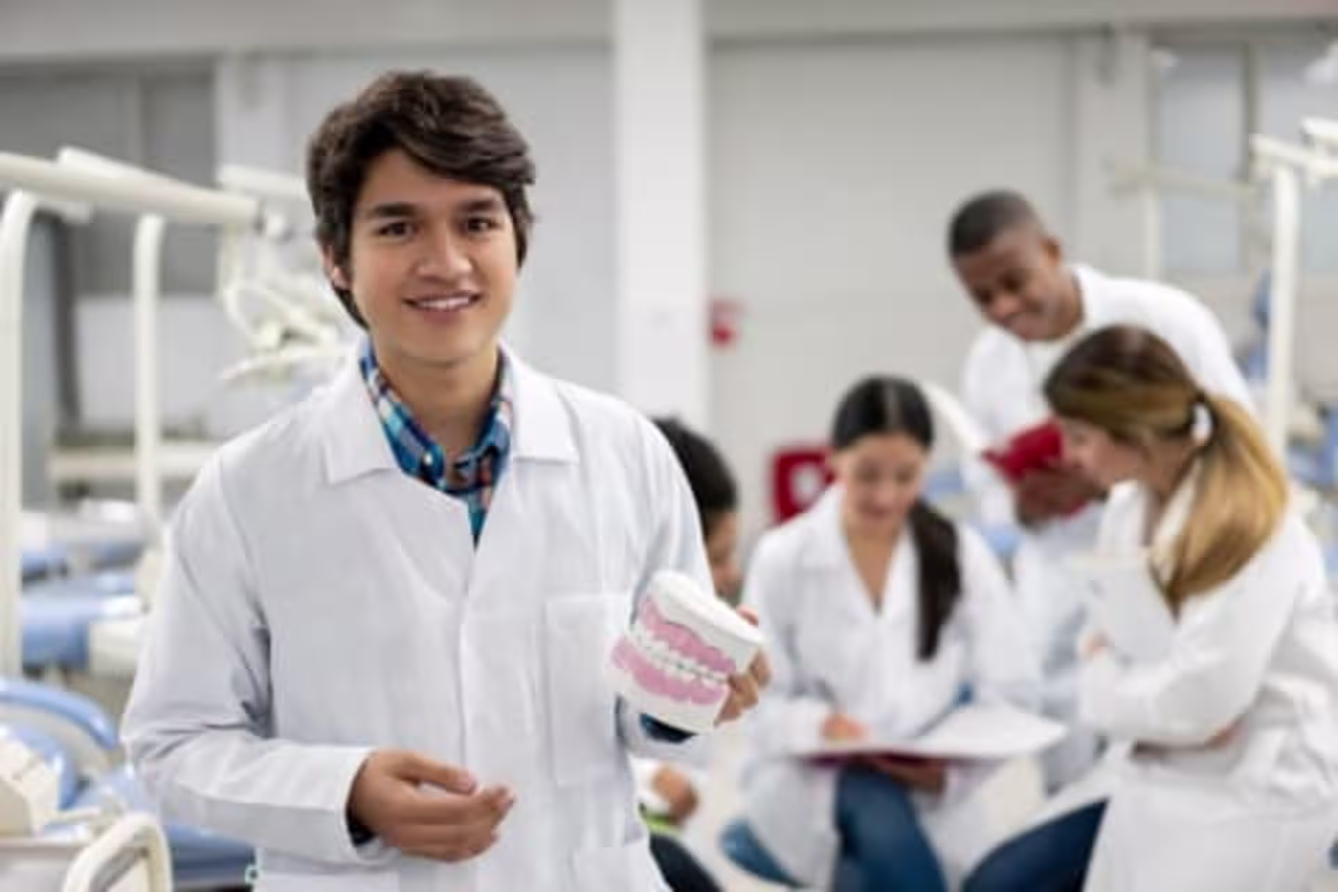A dental student wearing a lab coat smiles while holding a model of human teeth in a classroom setting, with other students studying together in the background.