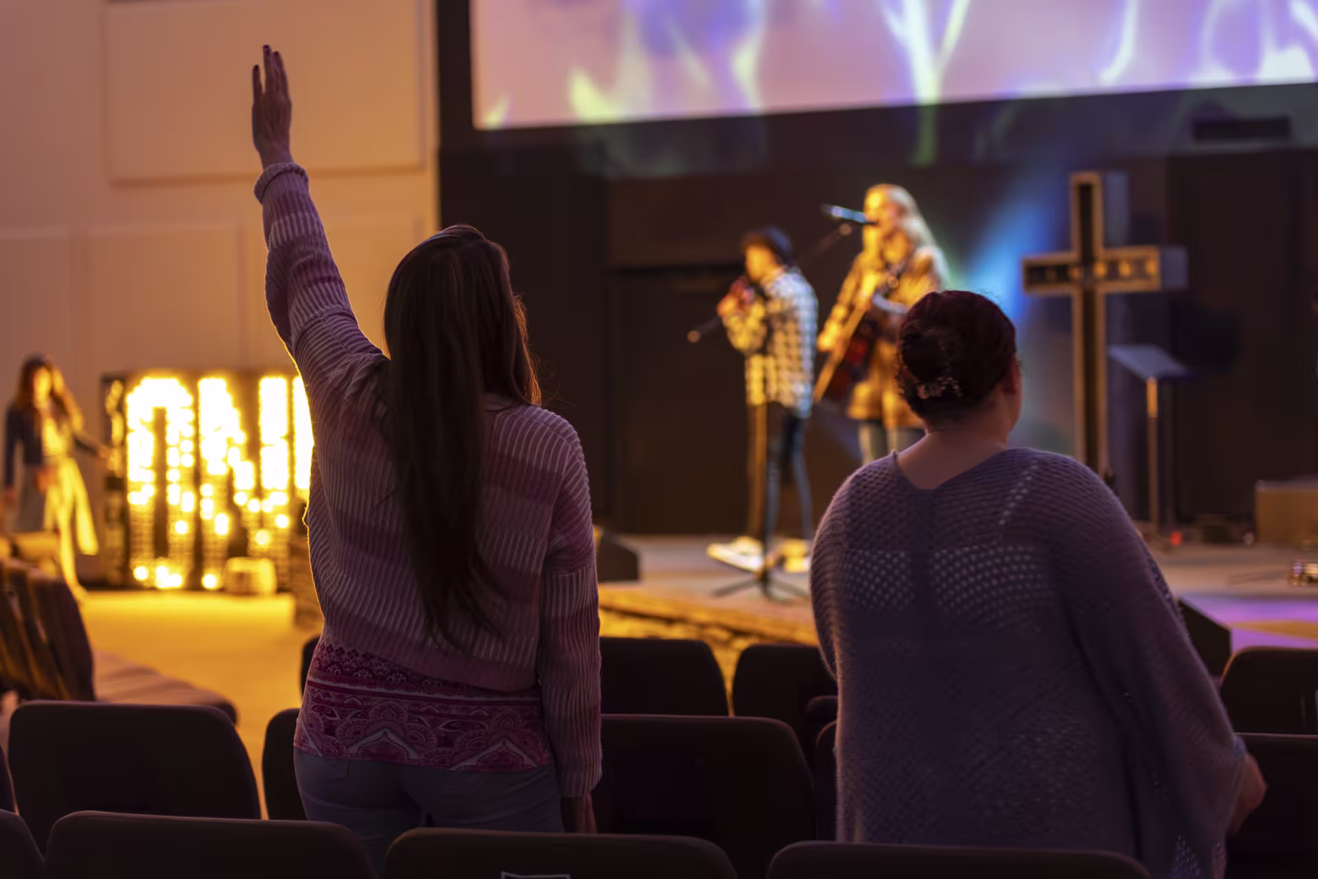 A group of people attending a worship service, raising their hands while musicians perform on stage. The setting reflects a vibrant urban ministry environment.