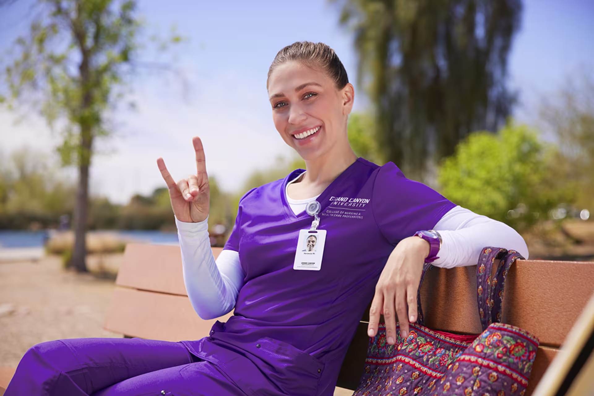 absn nursing student in purple scrubs sits on bench and gives lopes up sign