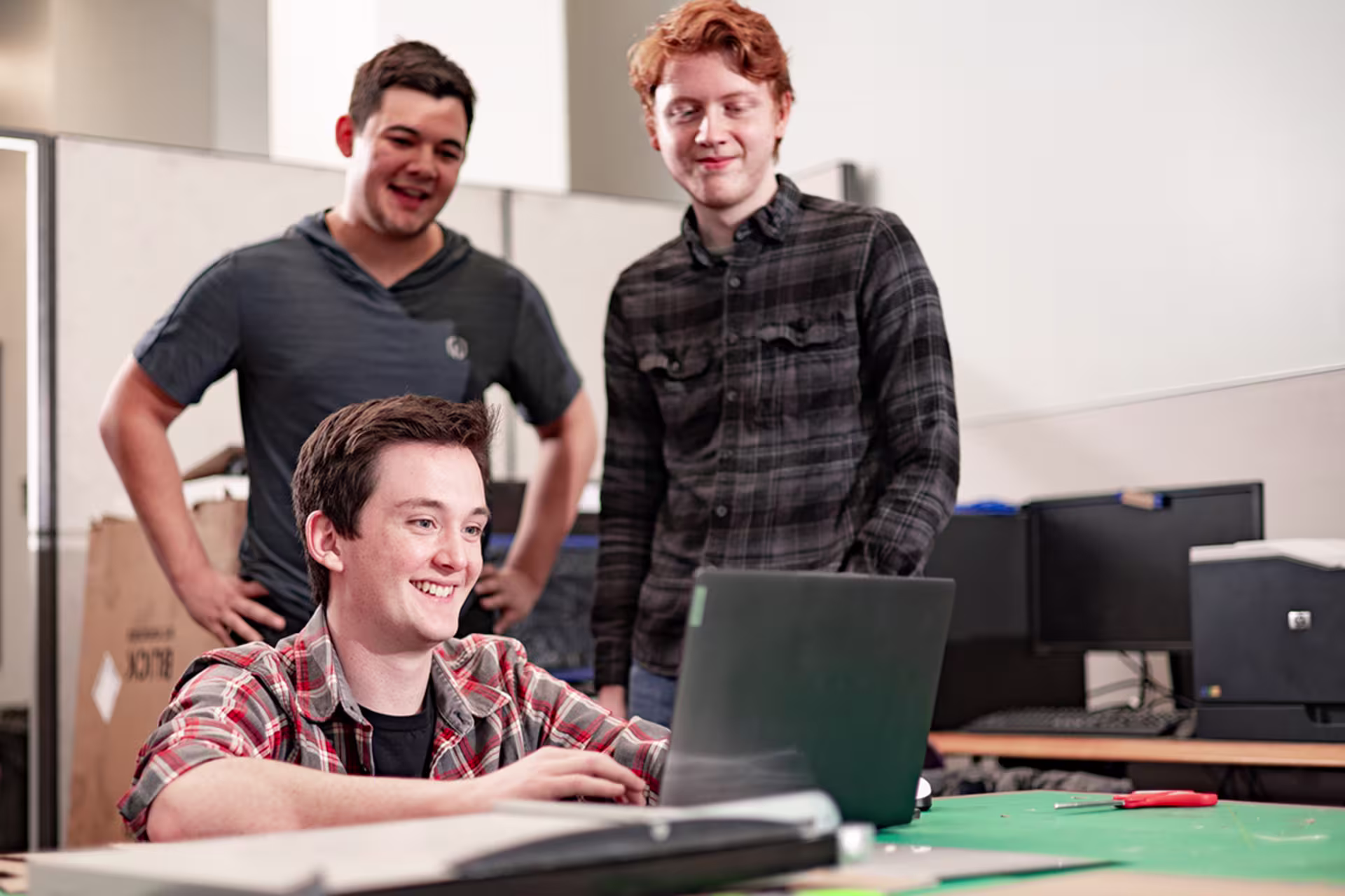 two men stand behind another on black laptop in engineering lab