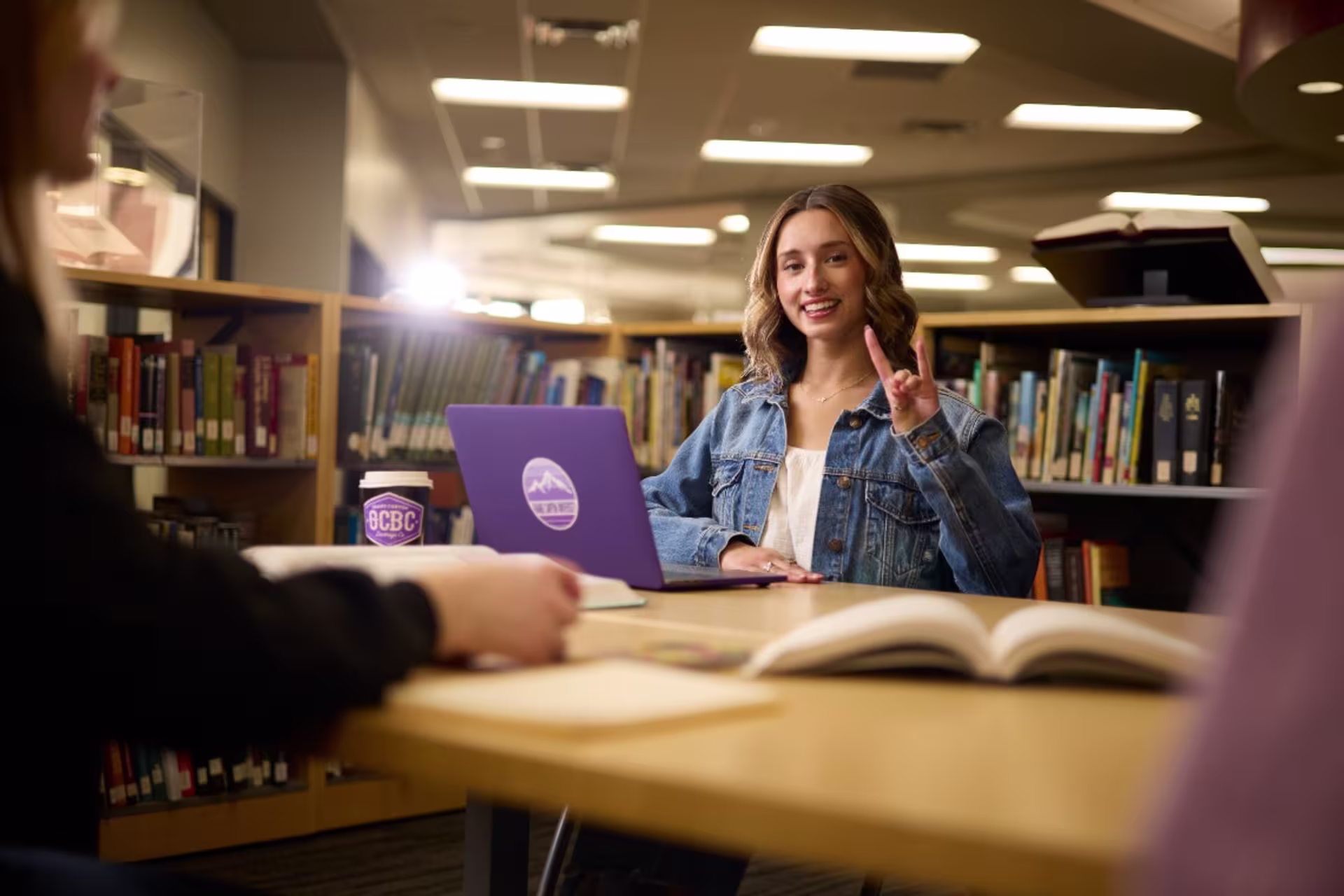 On campus student throwing lopes sign and sitting in library with laptop open with a study group.