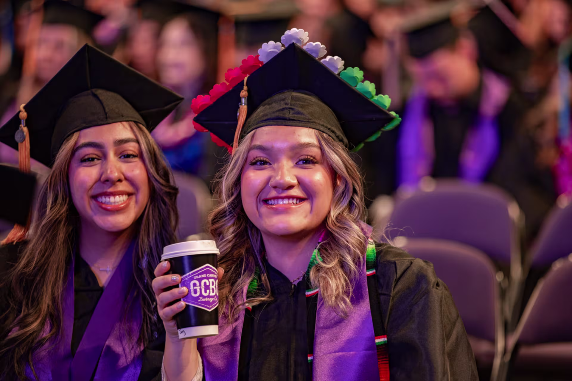 Female graduate smiles while holding up a GCBC cup at commencement with her friend next to her.