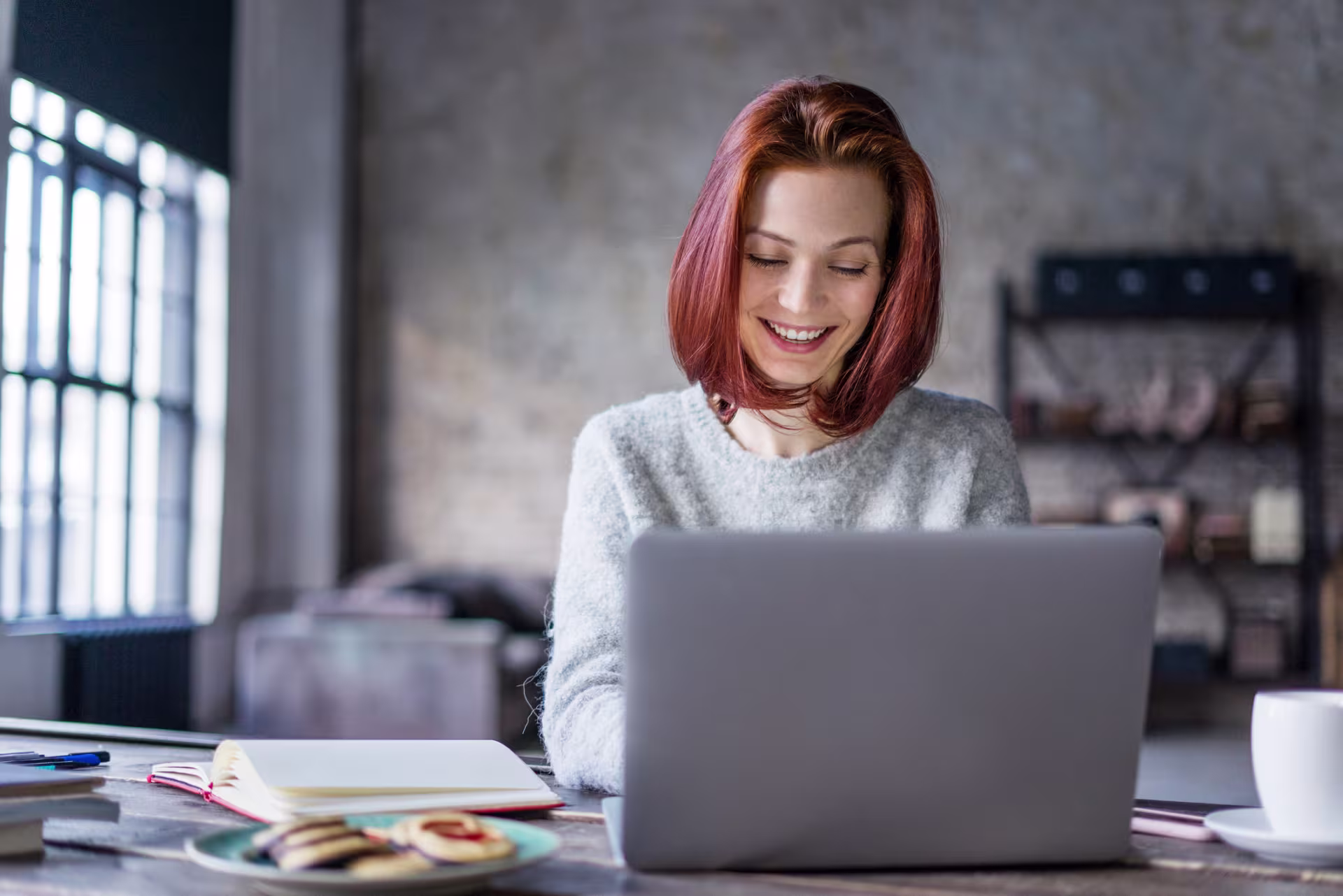 A red-haired person works on a laptop at a desk with a notebook and cookies, set in a room with industrial-style decor.